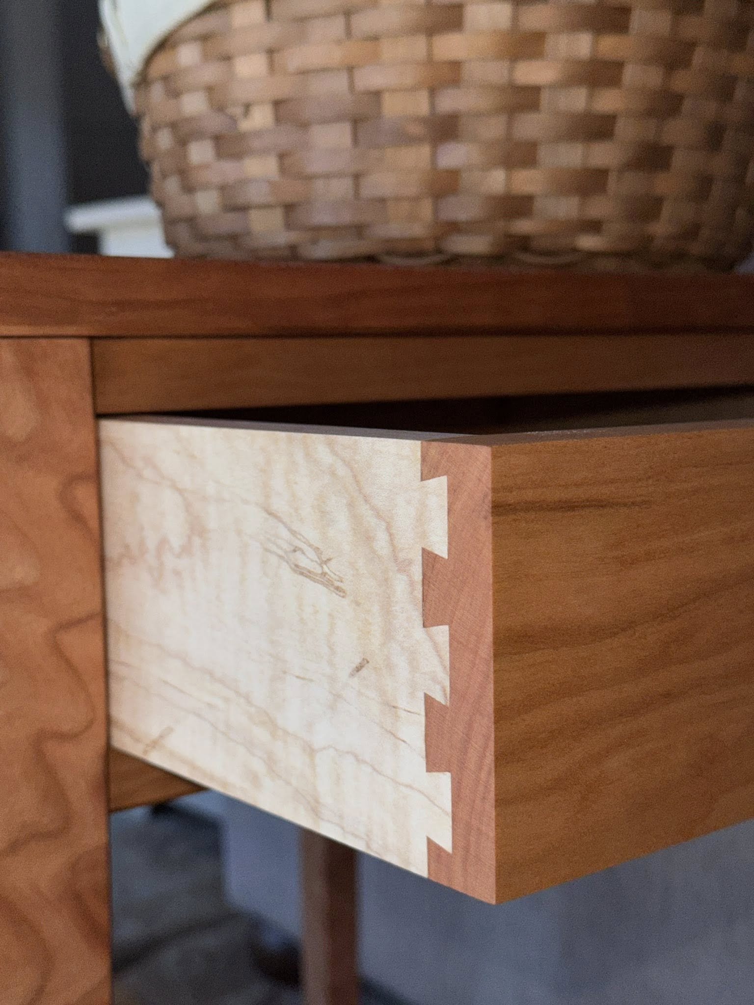 Close-up view of a cherry and maple wooden drawer displaying a dovetail joint, with a woven basket on top.