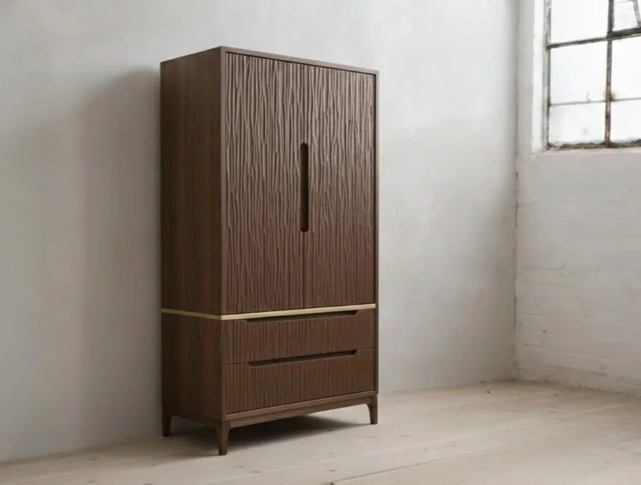 A tall wooden wardrobe with a vertical handle on the door, standing on four legs in a room with white walls, a large window with grid panes, and a light-colored floor.