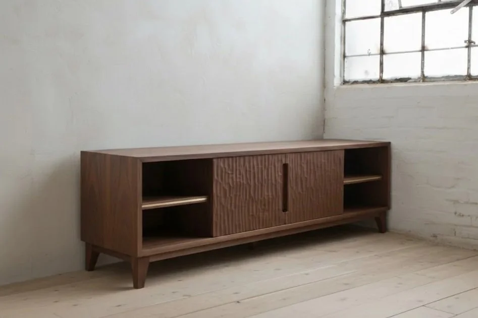 A wooden sideboard with open shelves and sliding doors in a minimalist room with white walls, a window, and light wooden flooring.