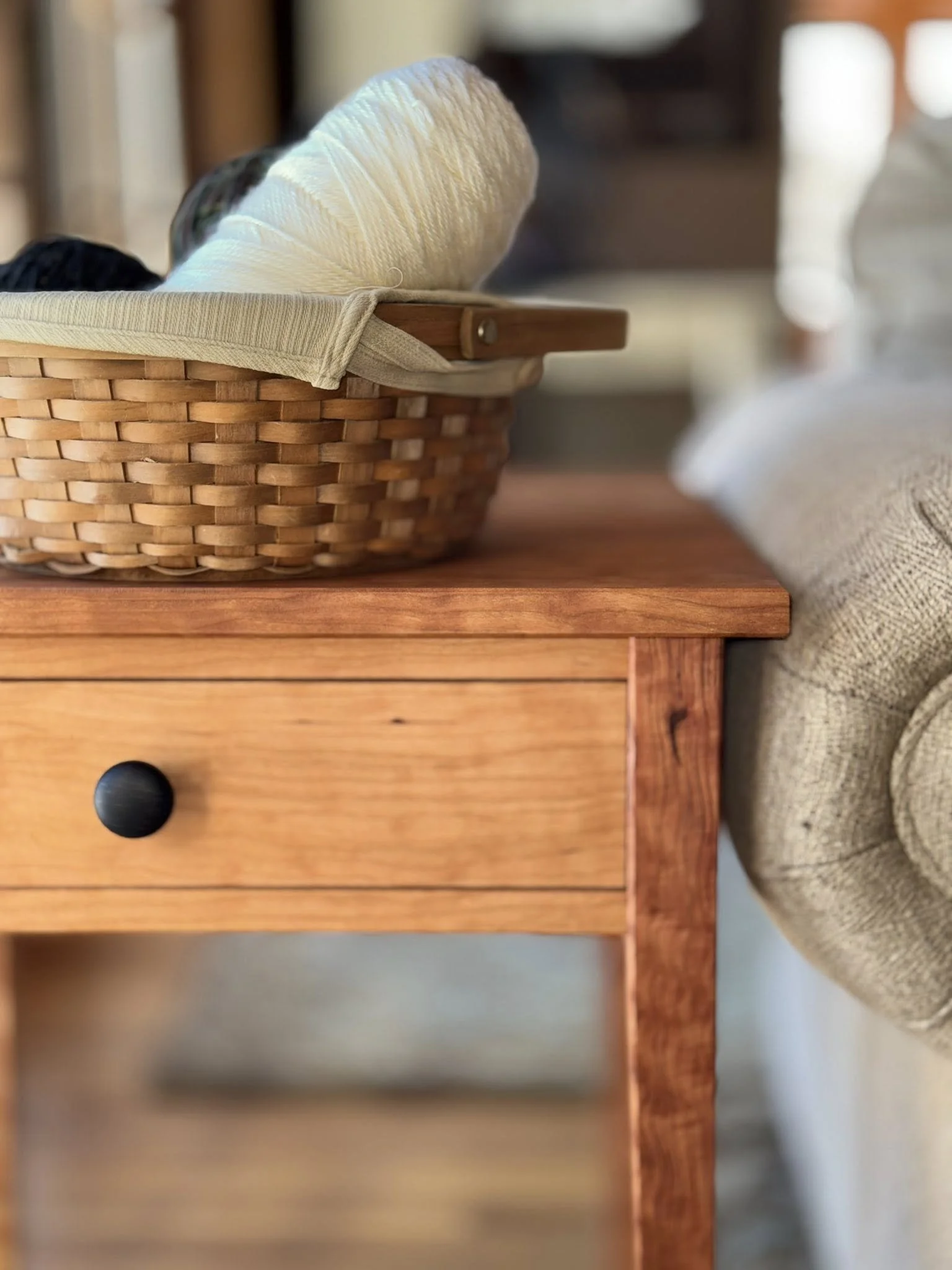 A wooden side table with a drawer, next to a beige fabric sofa, with a wicker basket containing yarn and knitting supplies on top.
