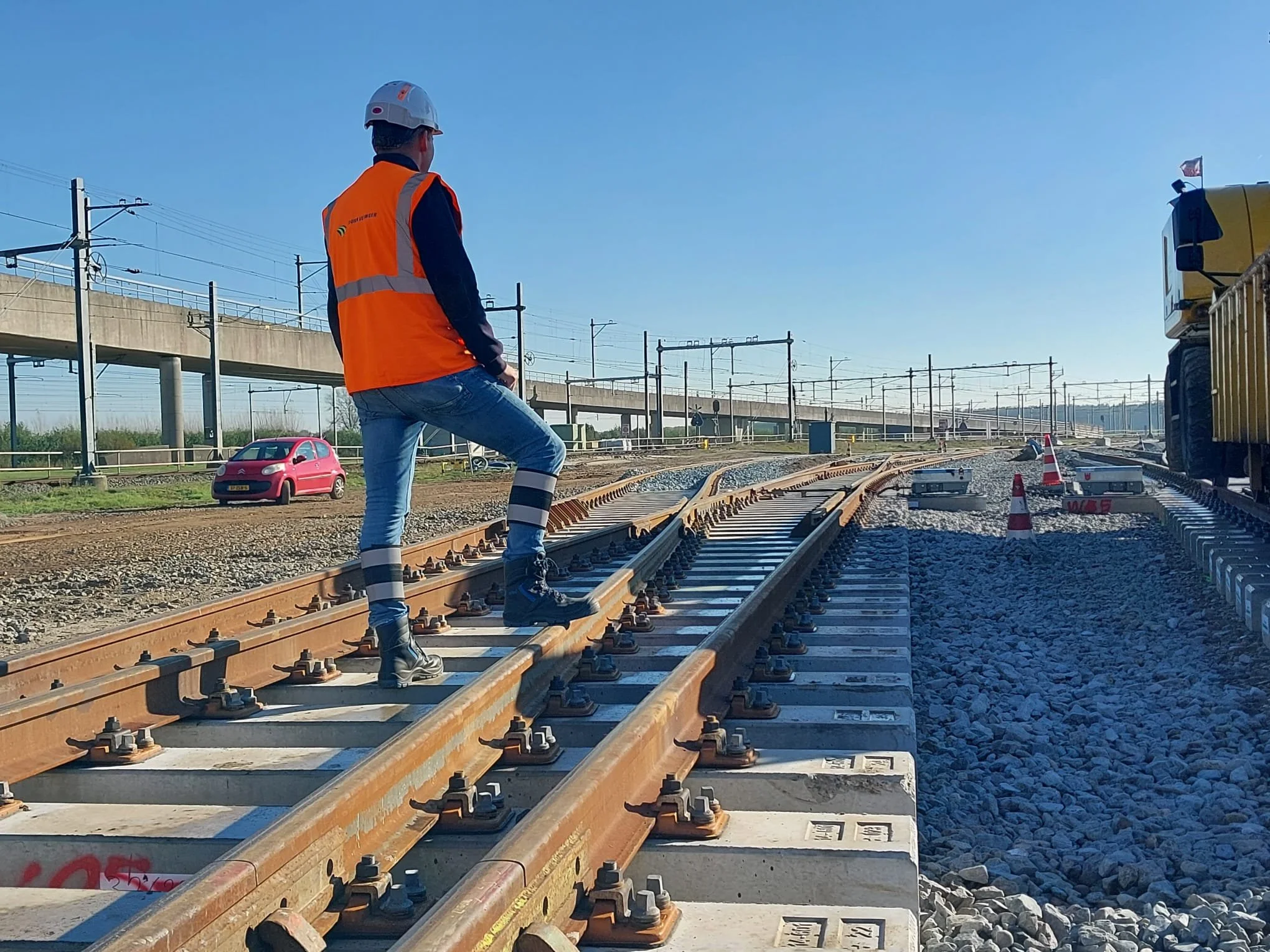 Arbeider in veiligheidsvest en helm die over spoorlijn loopt op een zonnige dag met een blauwe lucht, met een trein en auto op de achtergrond.