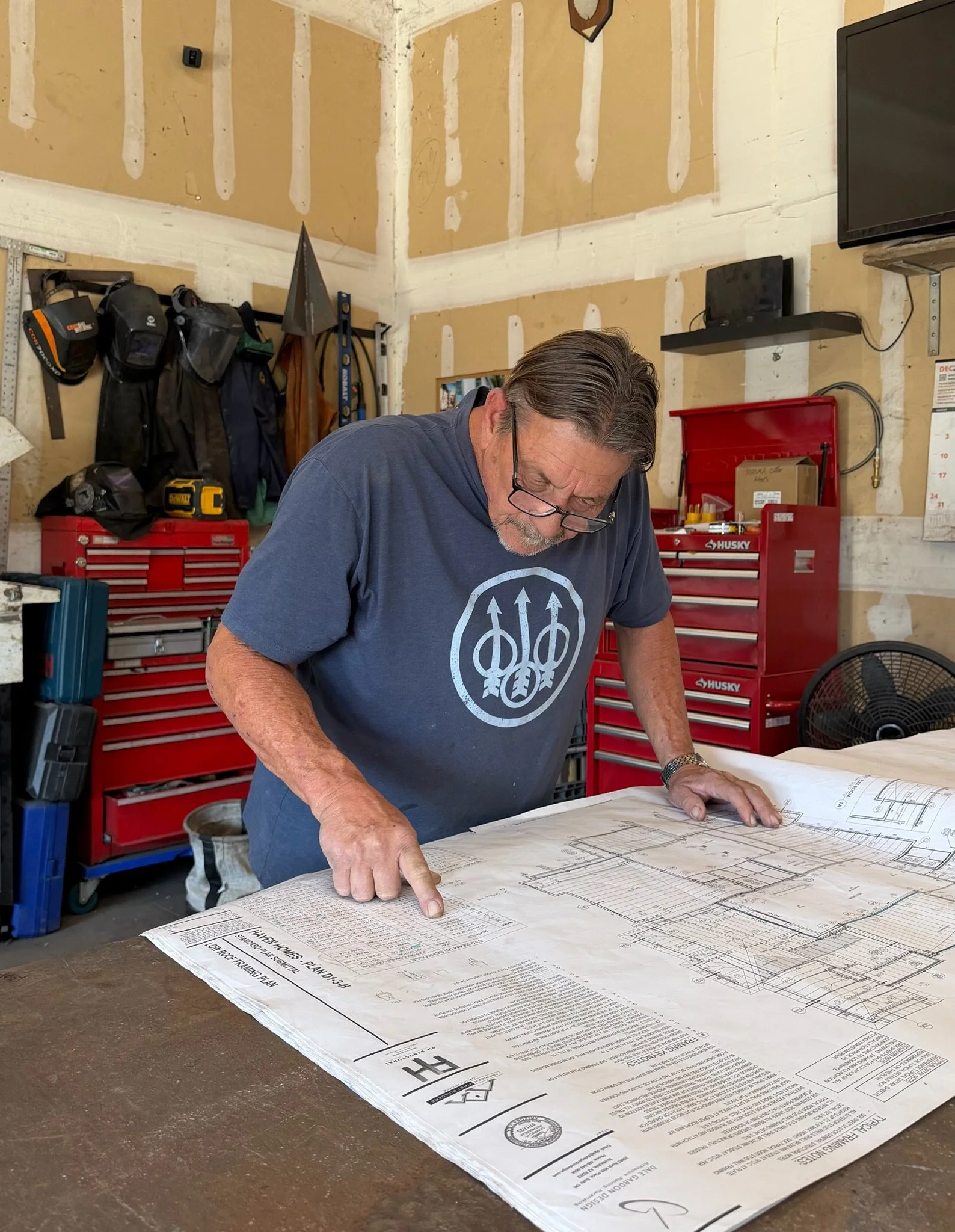 A man inside a workshop looking at and pointing to technical blueprints or architectural plans on a table.