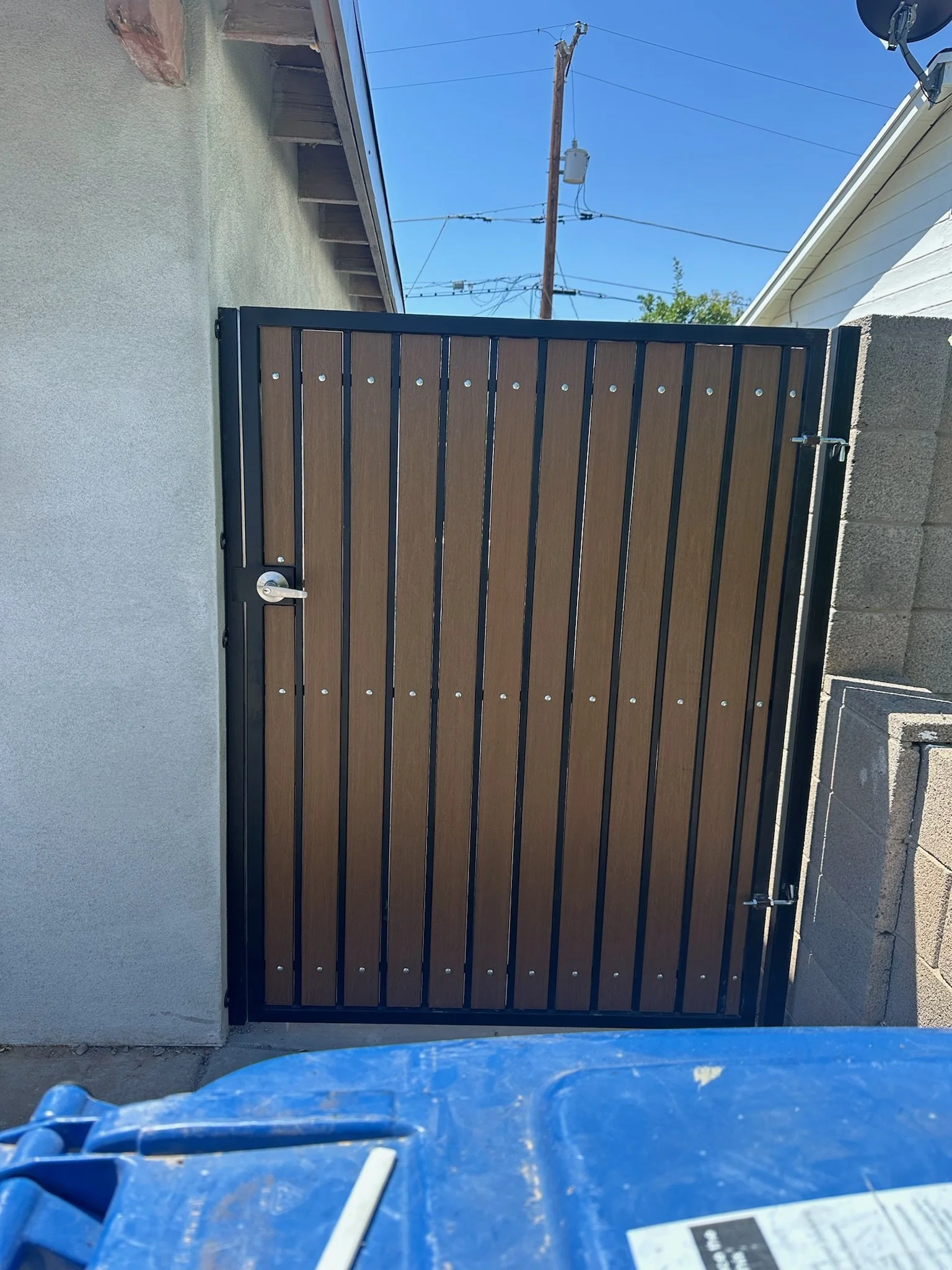 A wooden gate with vertical planks and black metal frame, installed between a light gray stucco wall and a concrete block wall, under a blue sky with utility poles and wires in the background.