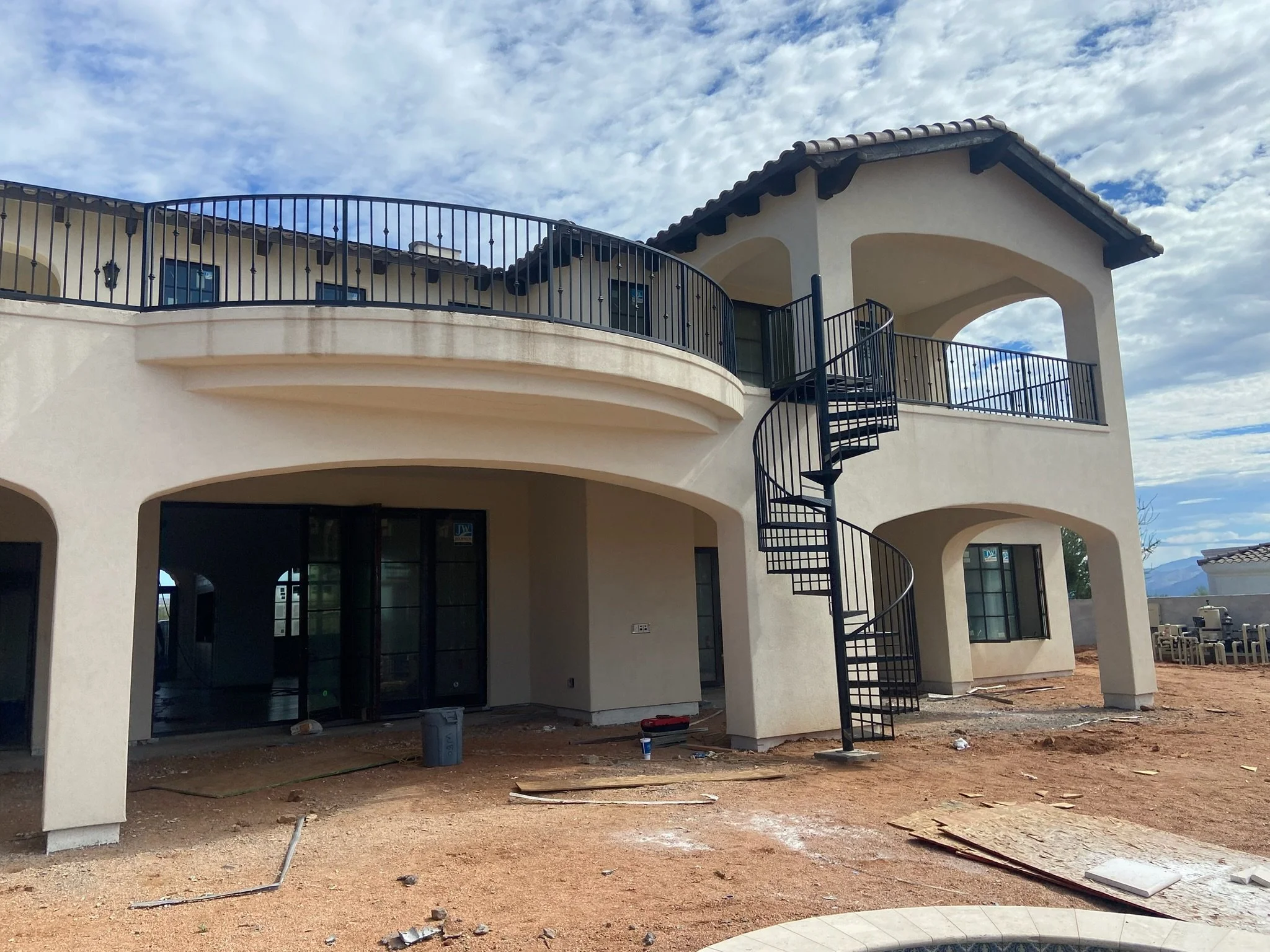 Newly constructed two-story house with beige stucco exterior, black metal railings, and a spiral staircase outside. The yard is unfinished with dirt and construction debris.