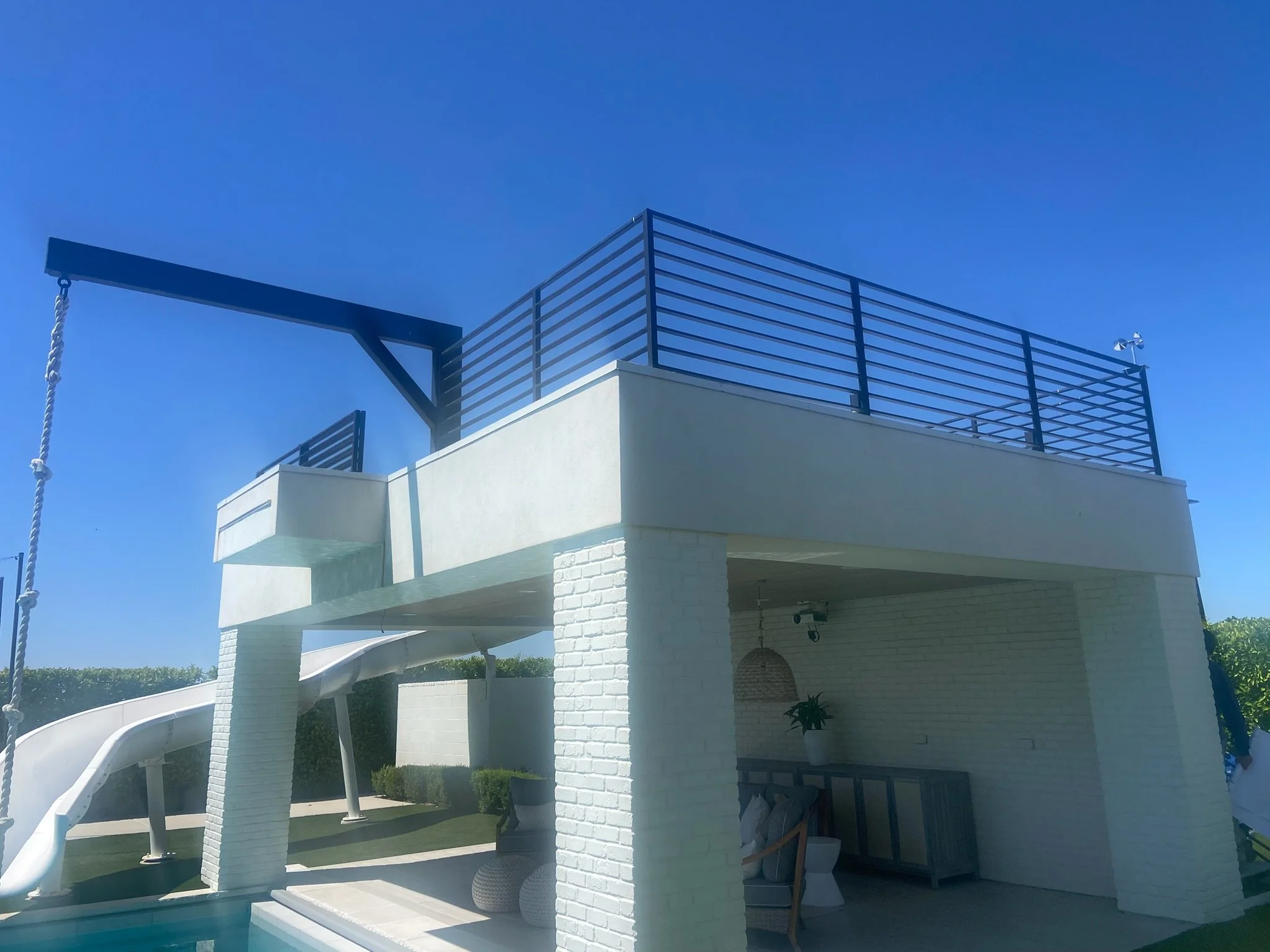 Modern two-story white house with outdoor slide, swing, and rooftop deck with black railing, under clear blue sky.