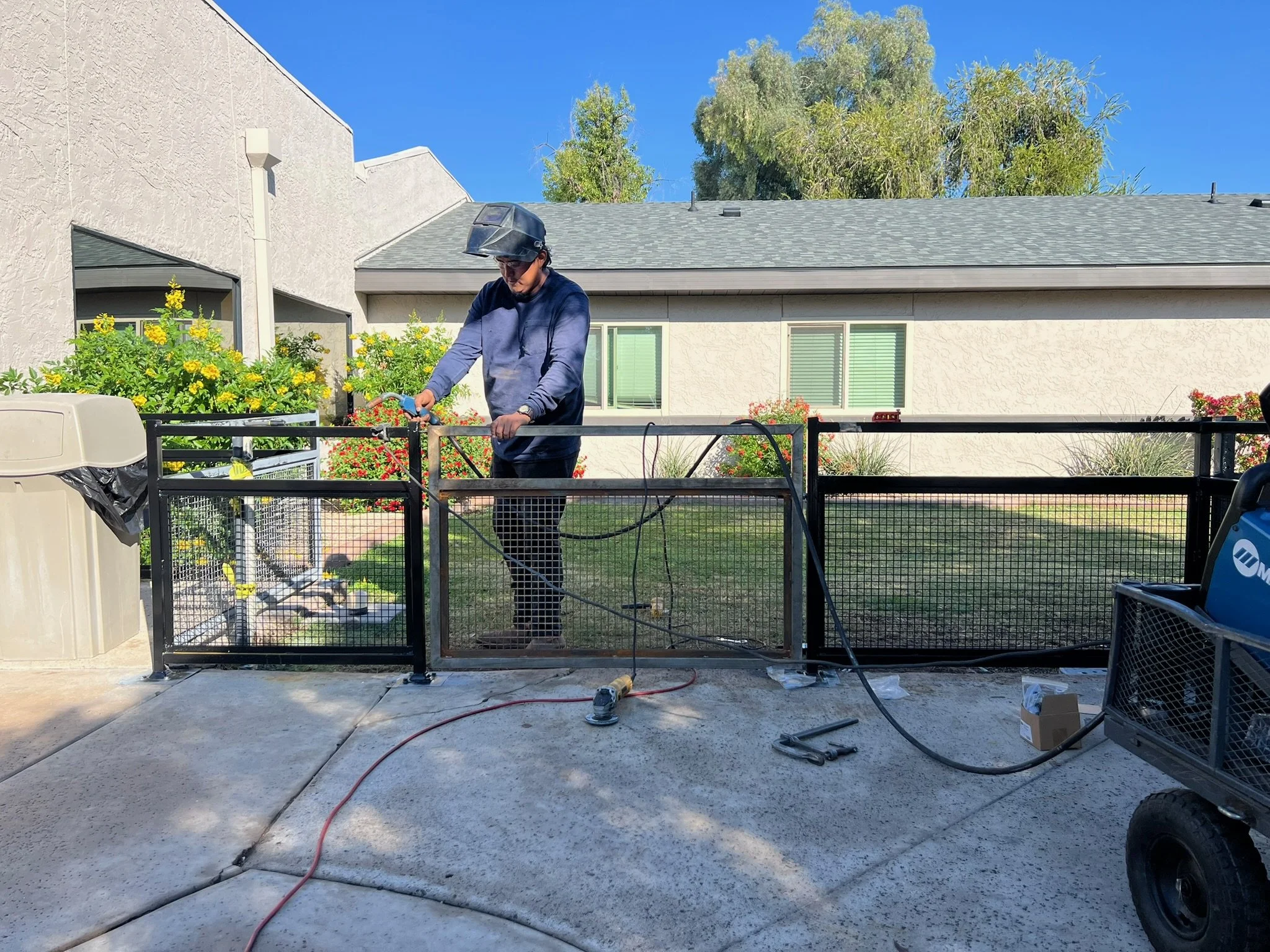 A person wearing a helmet and safety glasses is welding or grinding a black metal fence outside in a backyard. There are flowers and bushes behind the fence, a beige house with windows, a trash can, and a cart with equipment nearby. The sky is blue a