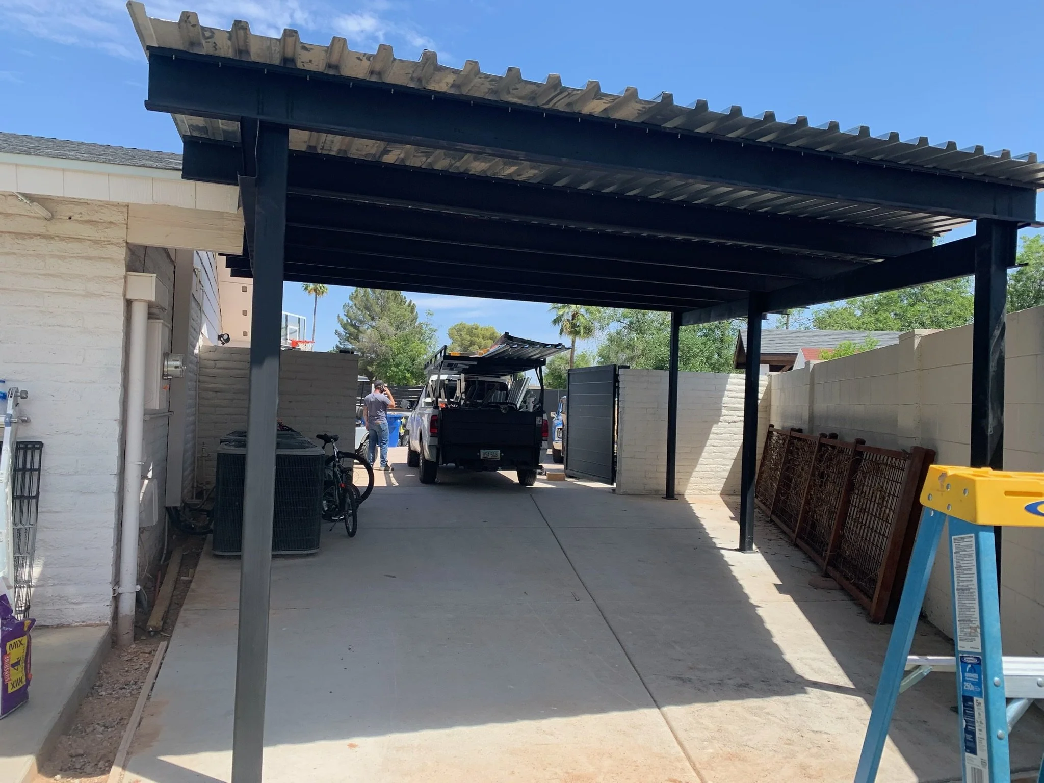 View of a carport with a black metal roof, adjacent to a white brick building, with a driveway leading to parked cars and a person, trees, and a blue sky in the background.