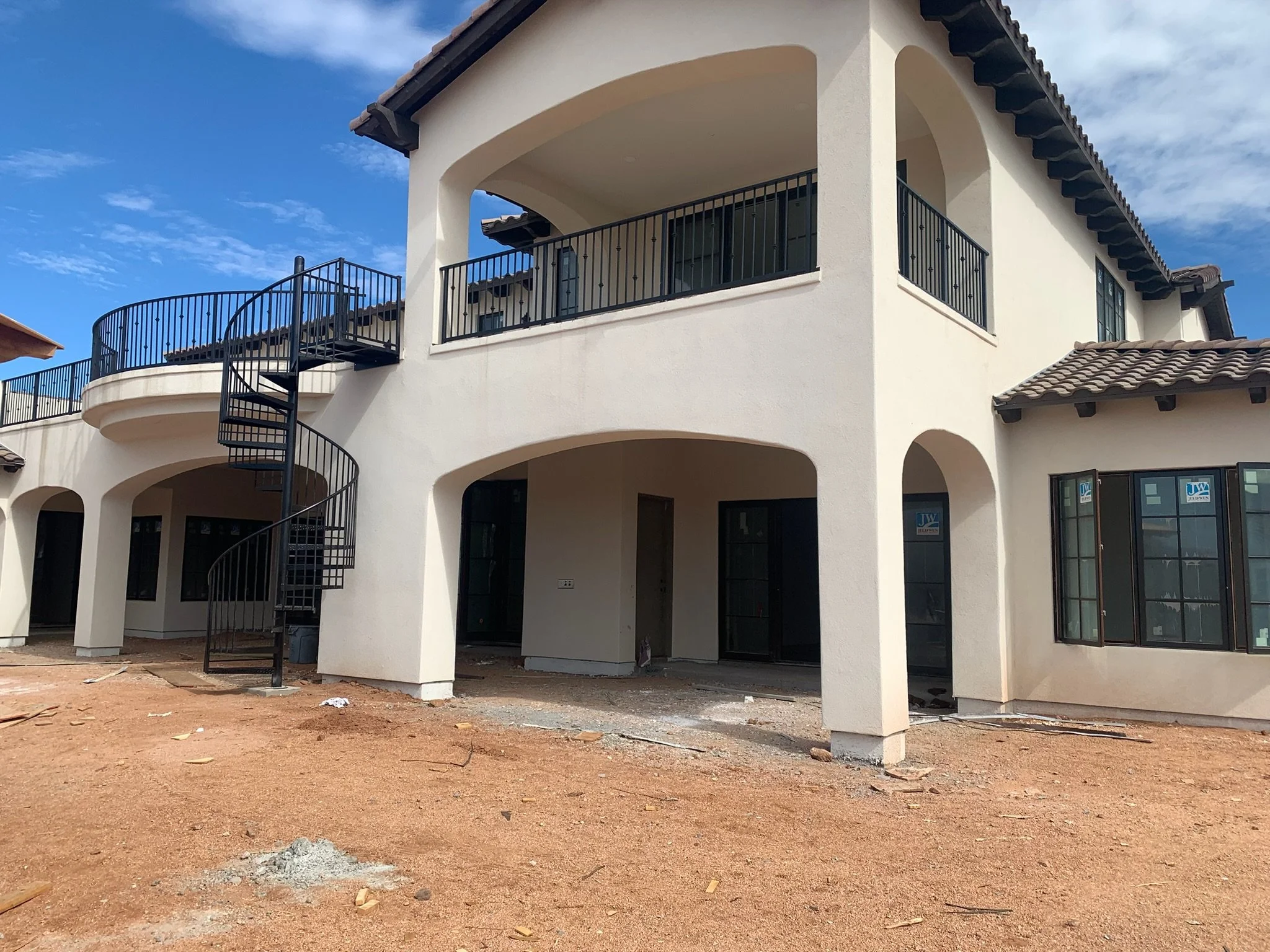 The image shows a modern, multi-story house under construction with white stucco exterior walls, large windows, and a black metal spiral staircase on the left side leading to a balcony. The ground is bare dirt.