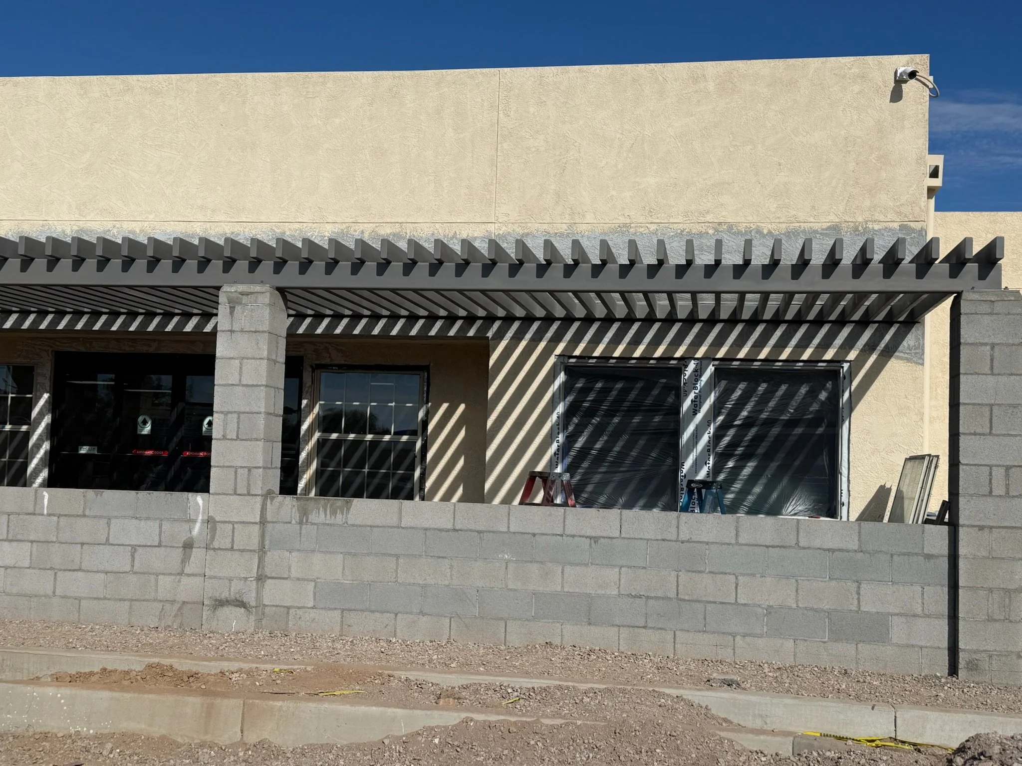 Construction site of a building with a beige exterior wall, a black pergola on the patio, and a cinder block wall in front. There are two windows, one covered with plastic, and construction tools and materials on the patio.
