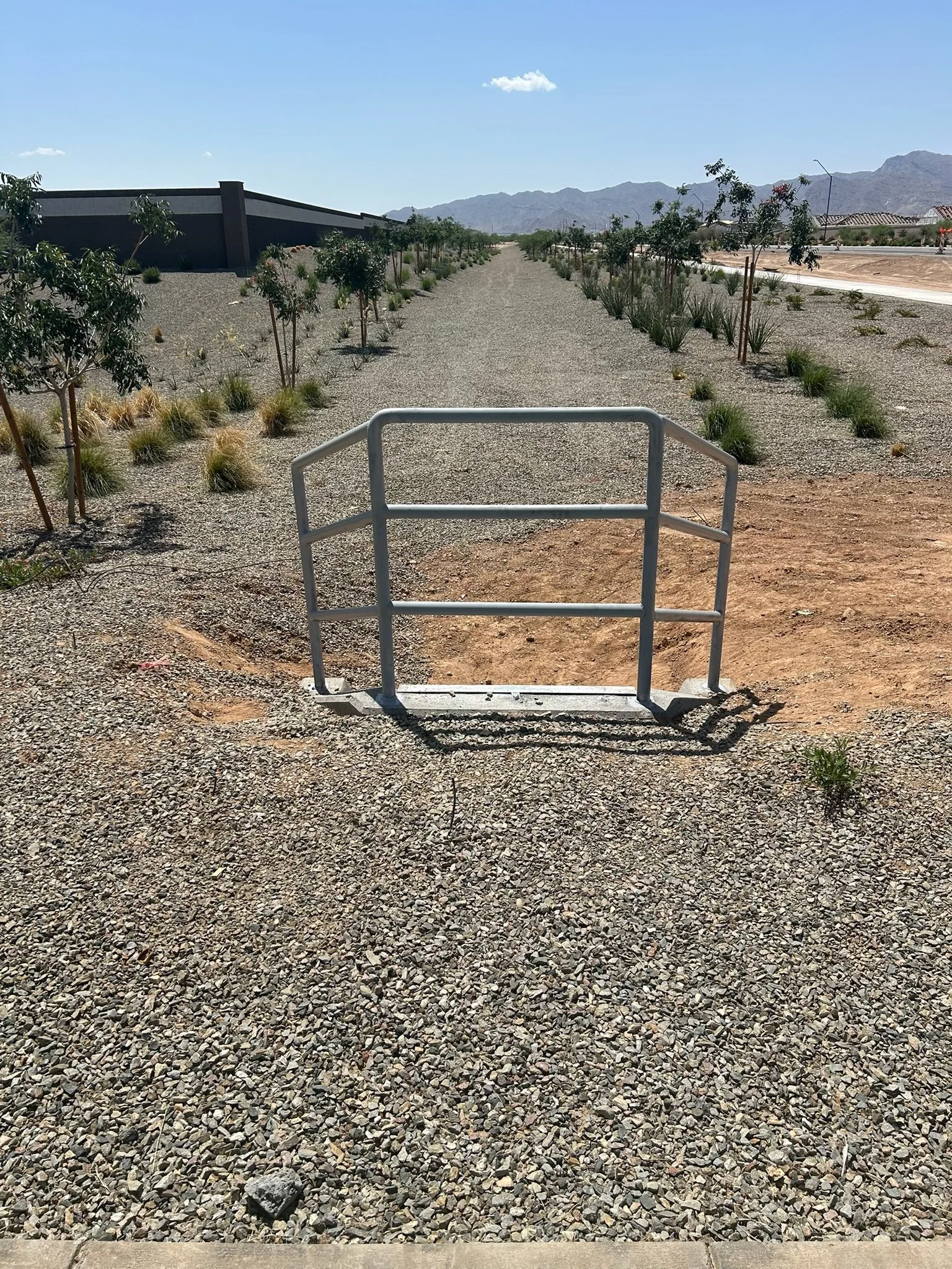 A metal barrier on a gravel pathway in an arid landscape, with small trees and bushes along the sides and mountains in the background.