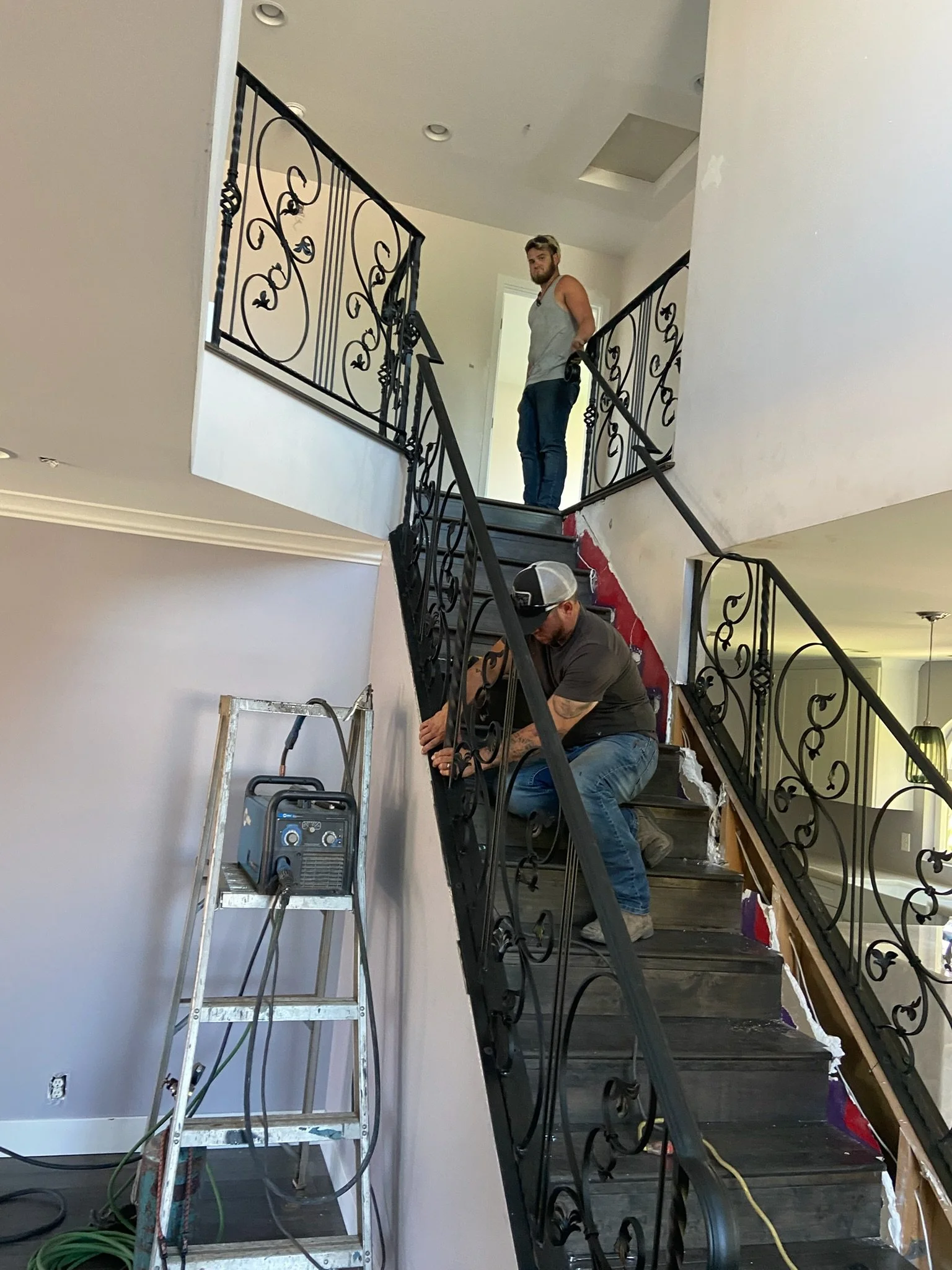 Two men working on a staircase in a house, one kneeling and using tools on the black metal railing, the other standing at the top of the stairs observing. There is a step ladder with equipment on it beside the wall.
