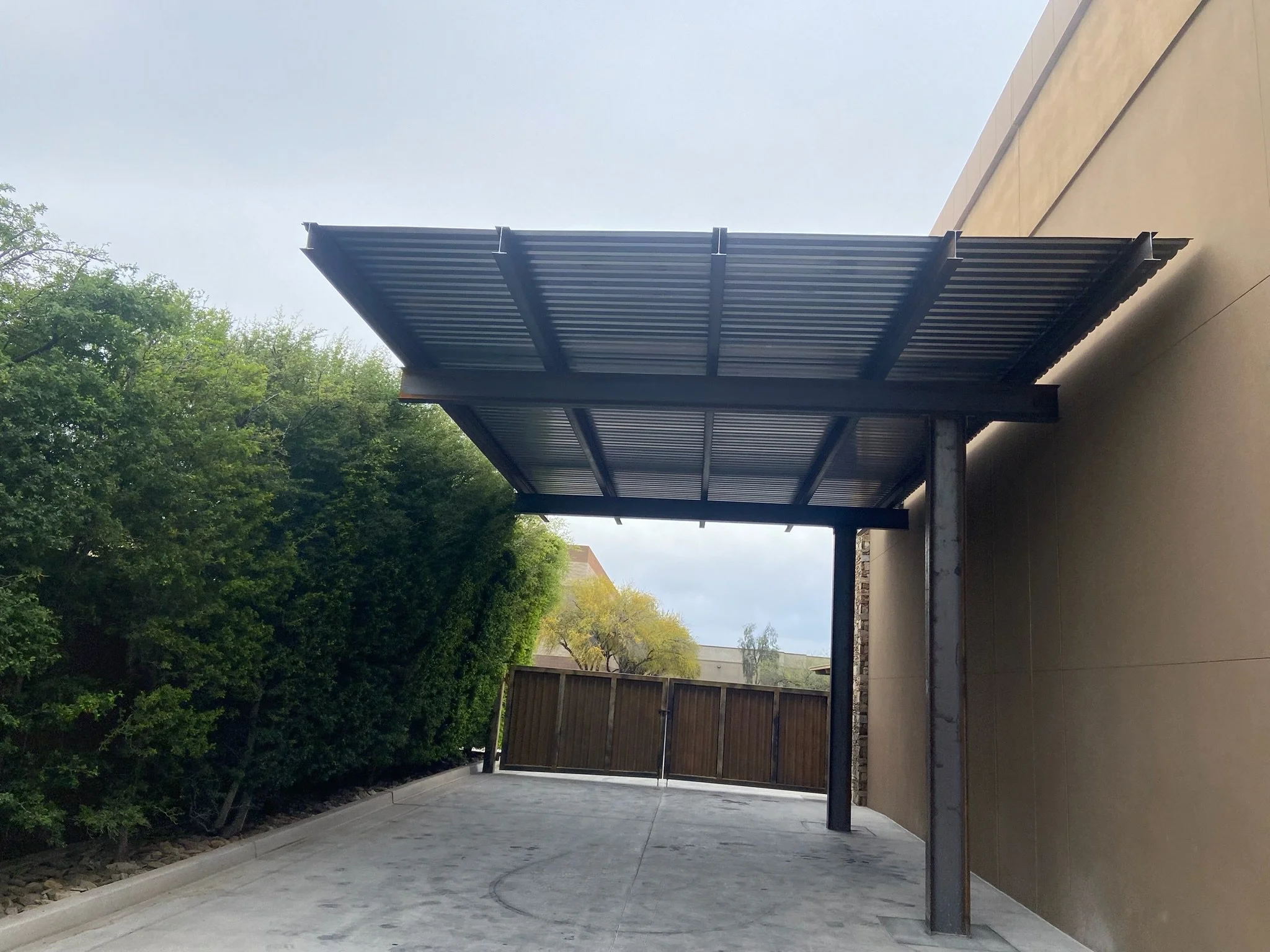 Modern metal awning attached to a building with trees and a wooden gate in the background.