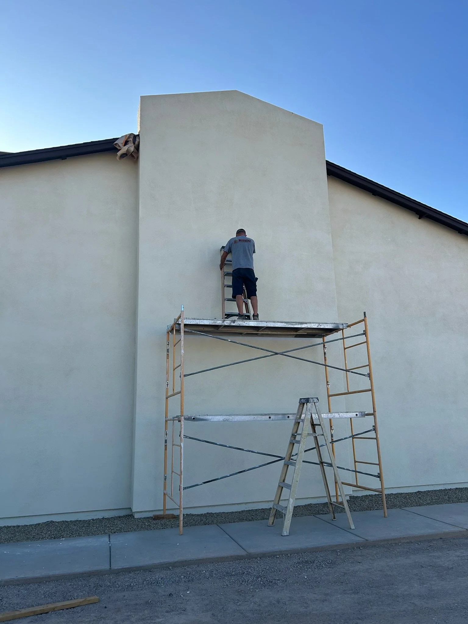 A person standing on scaffolding, painting the exterior wall of a building under a clear blue sky.