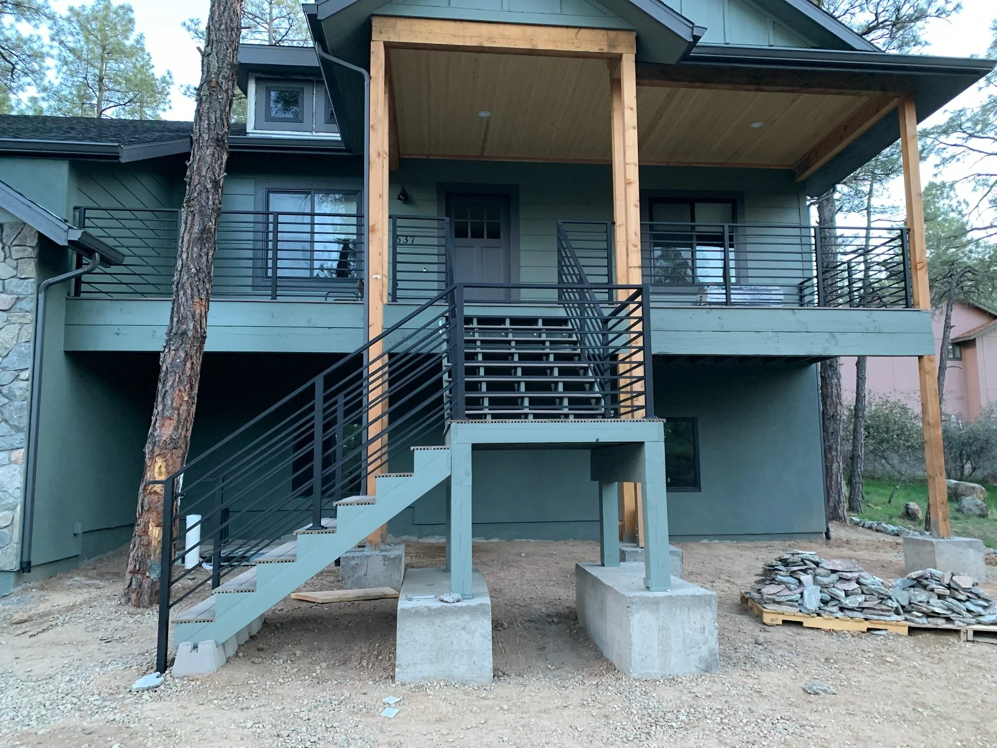 A two-story house under construction with a green exterior, a covered front porch with wooden beams, and black metal railings. The front stairs are concrete with black metal handrails. The ground is dirt, and there are piles of rocks nearby.