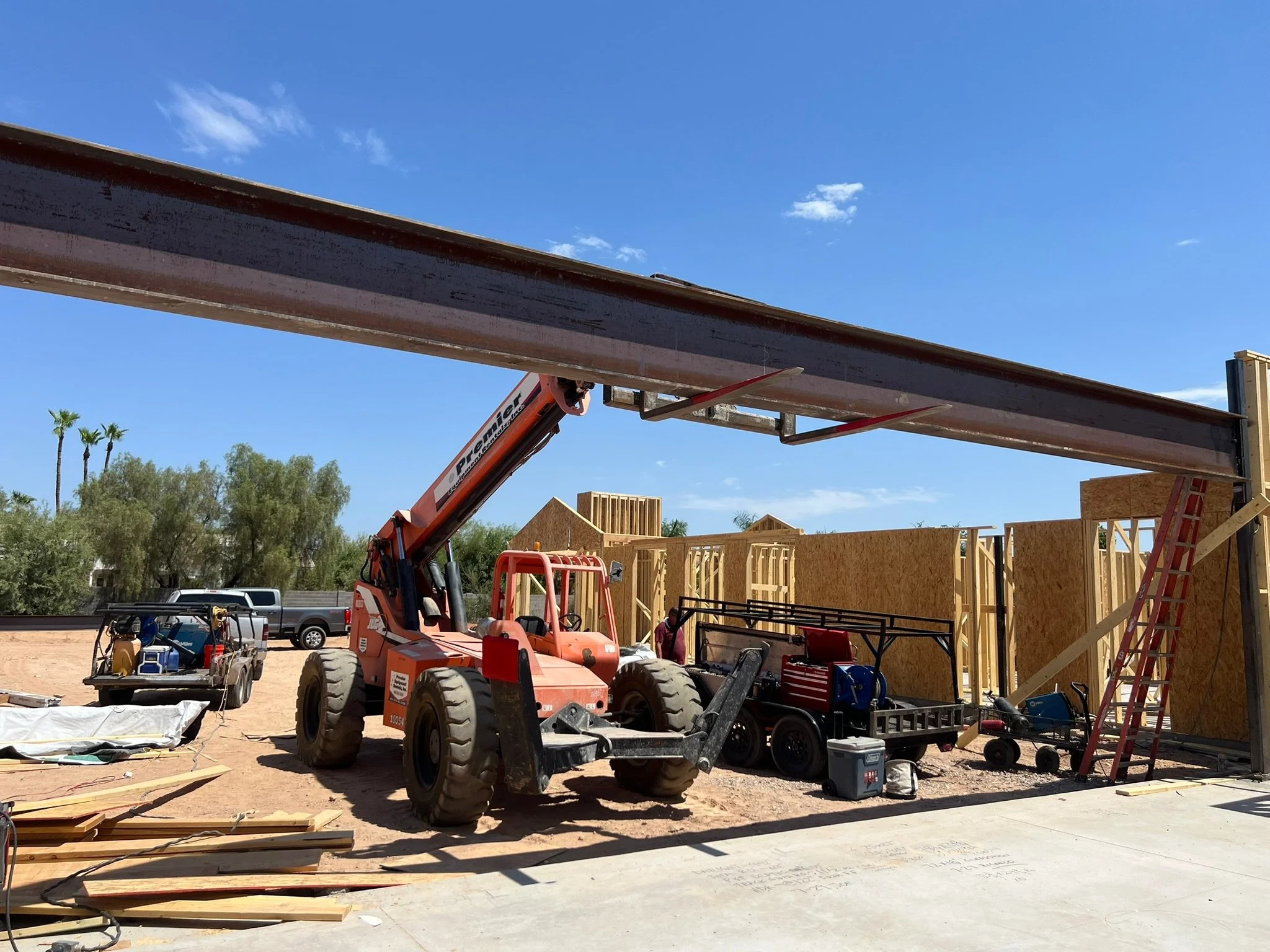 Construction site with a red crane lifting a steel beam, wooden framing of a building under construction, and construction tools and materials on the ground.
