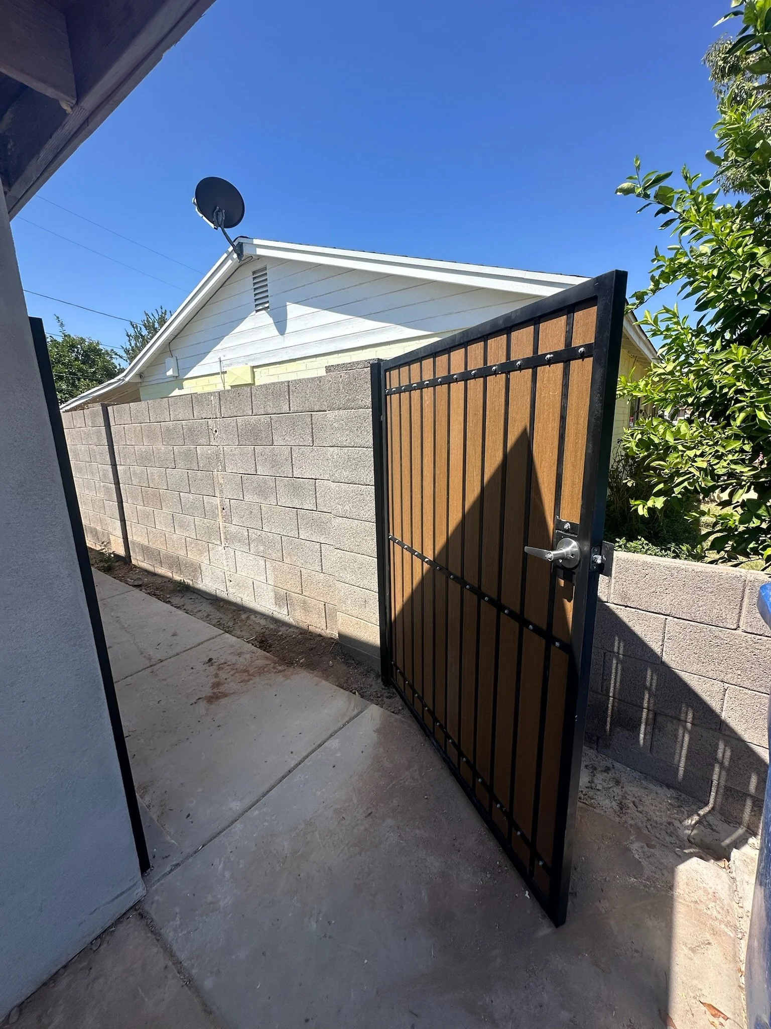 Open metal gate with wooden panels leading to backyard with concrete patio, brick wall, house with white siding, satellite dish, and blue sky.