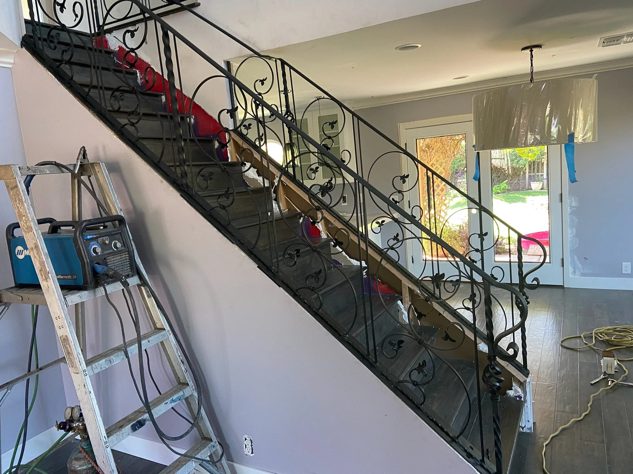 Interior of a house under renovation with a staircase, a ladder with a welding machine, and a large window door leading outside to a garden.
