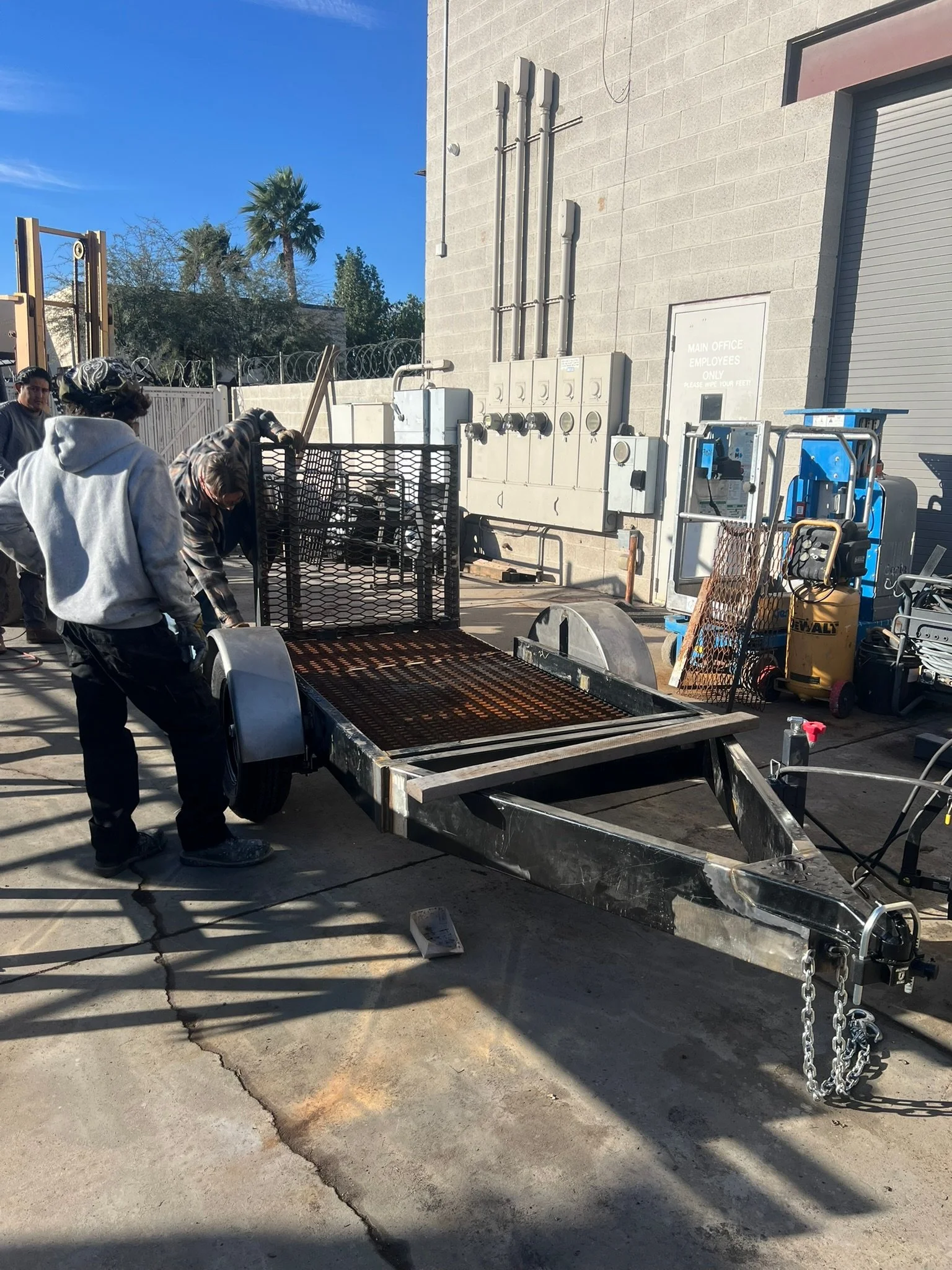 Workers install or repair a metal utility trailer with a grated ramp, situated on a concrete surface outside a building.