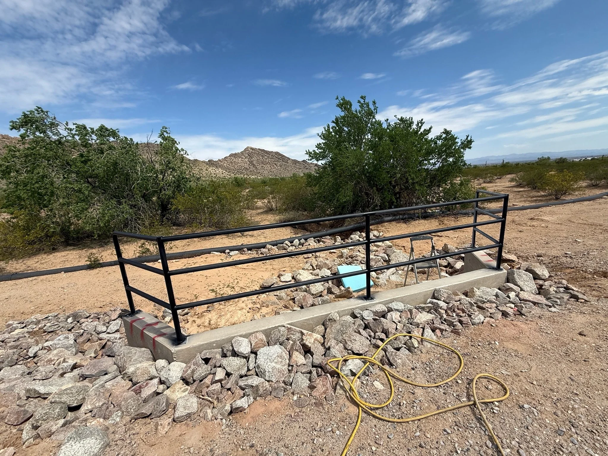 A newly constructed concrete ramp with black metal railings in a desert landscape with sparse bushes, a small hill, and mountains in the background. An orange extension cord lies on the ground in front of the ramp.