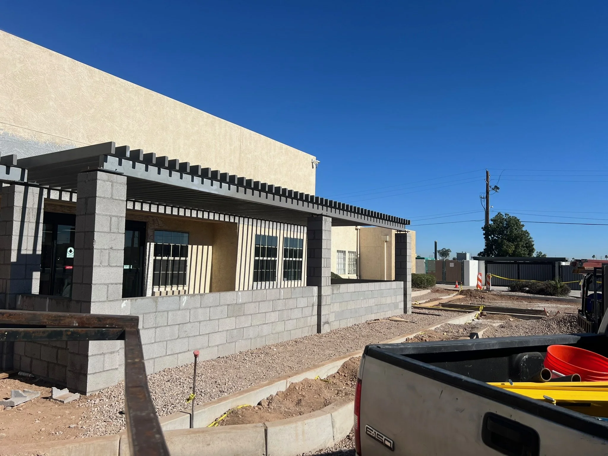 Construction site showing a building with a partially completed patio or porch area, construction materials, and equipment in the foreground, with blue sky and some trees in the background.