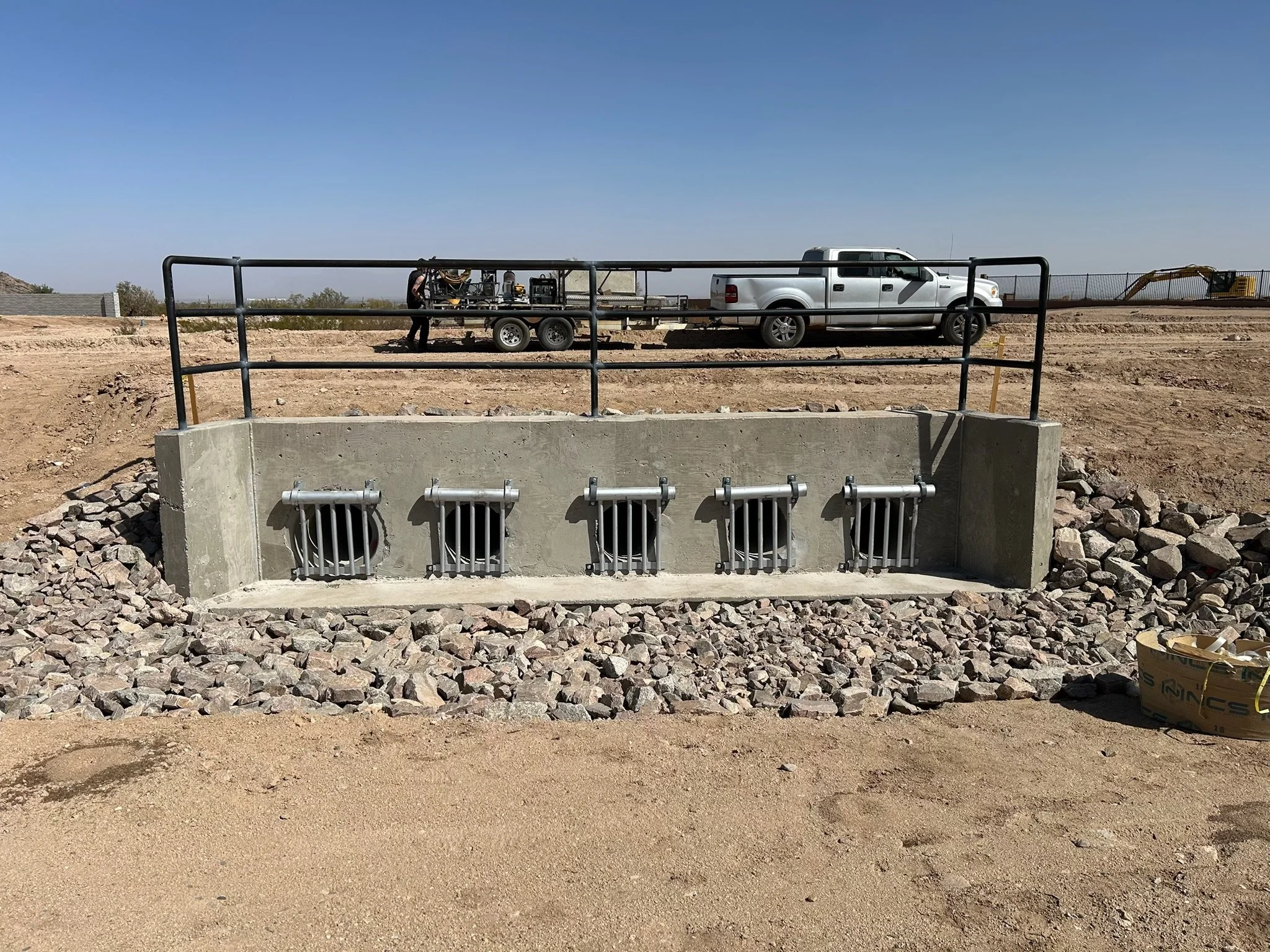 Underground utilities vault with four locked metal hatches, surrounded by rocks, at a construction site with a pickup truck, trailer, and construction equipment in the background on a clear day.