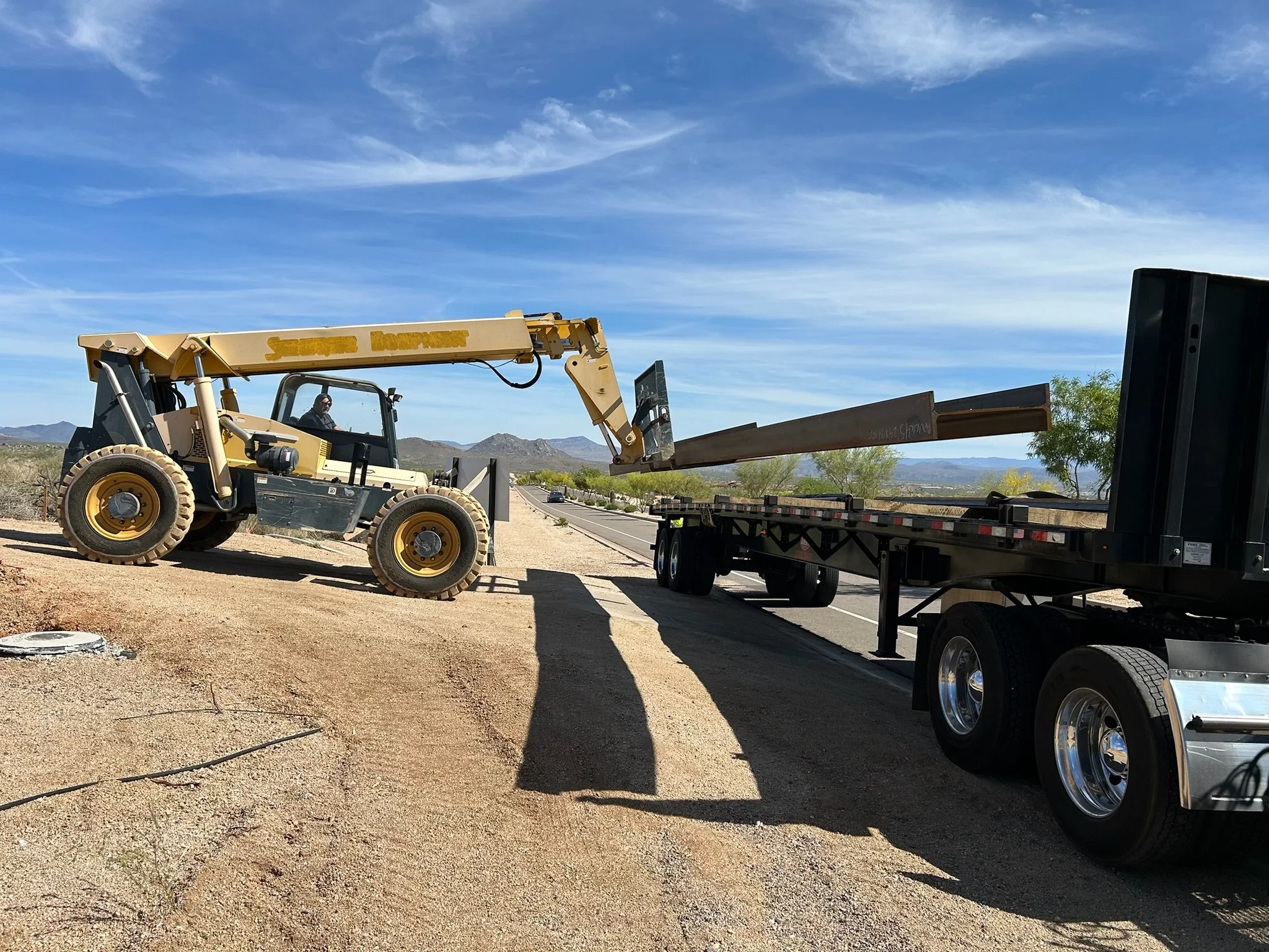 A bulldozer loading long metal beams onto a flatbed truck on a desert road with mountains and trees in the background.