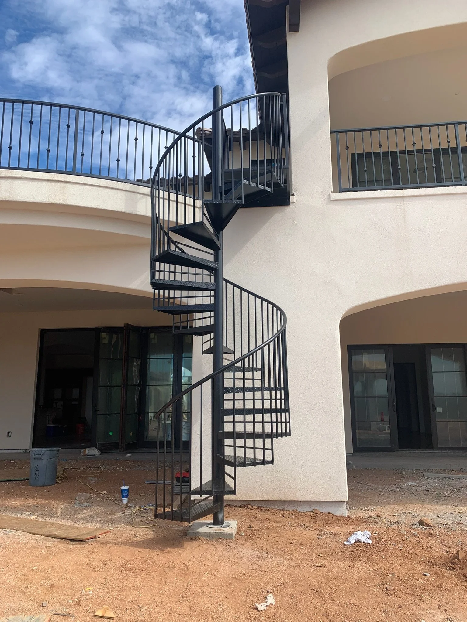 Black metal spiral staircase outside a white building under construction, with dirt ground, construction debris, and large windows.