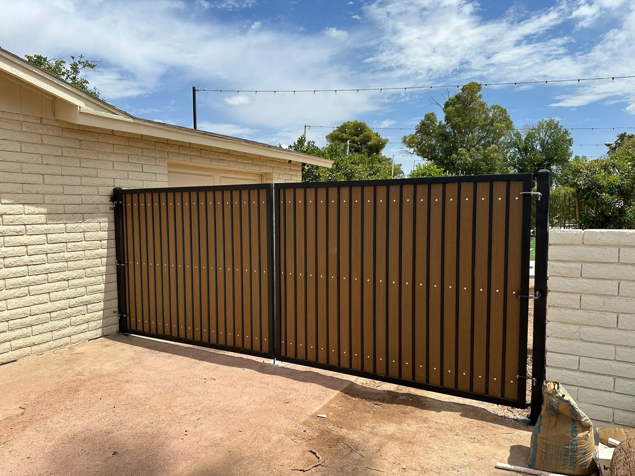 A black metal gate with wooden slats installed between a beige brick wall and a white brick wall, under a partly cloudy blue sky.