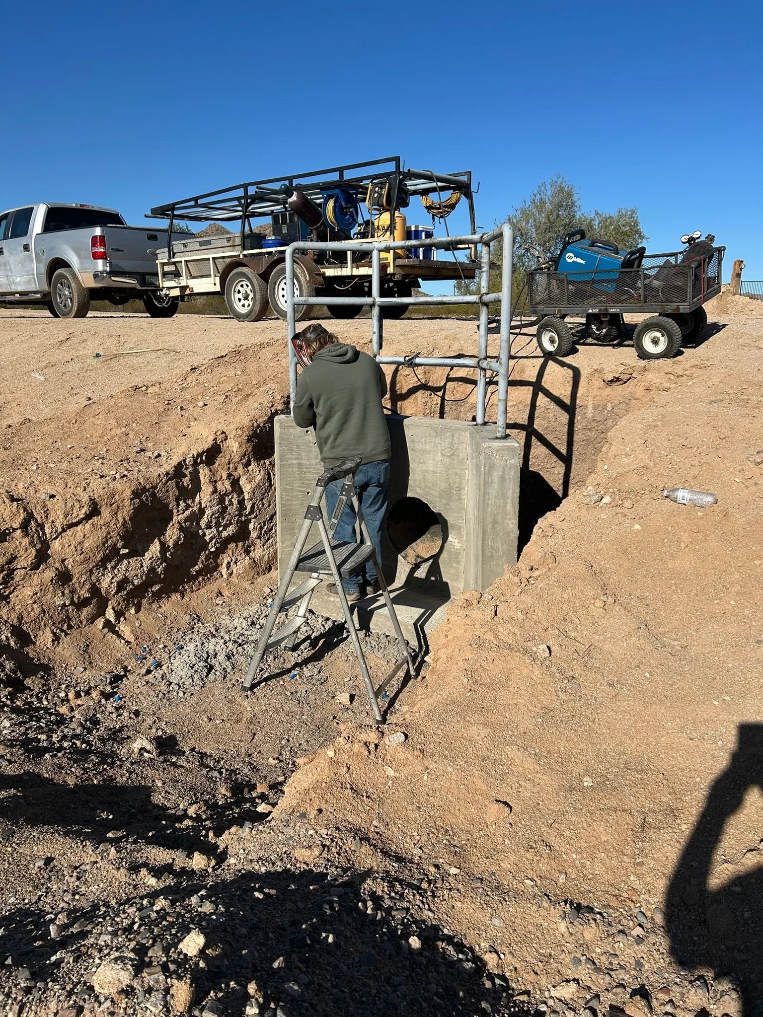 Worker installing or inspecting a concrete pipe under a road, with construction equipment and vehicles in the background.