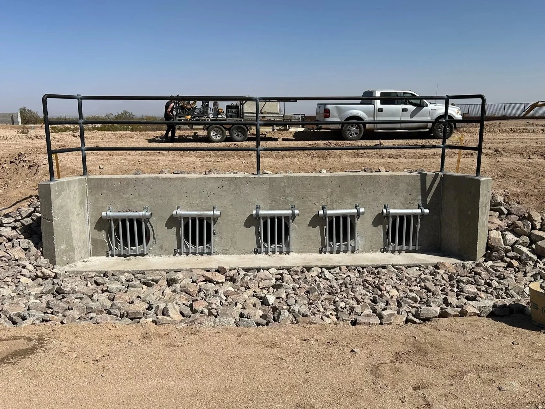 Concrete structure with four small barred openings, equipped with metal grates, surrounded by rocks, and a black metal railing on top. In the background, a white pickup truck is parked, and a trailer attached to it carries equipment. The scene is outdoors under a clear blue sky.