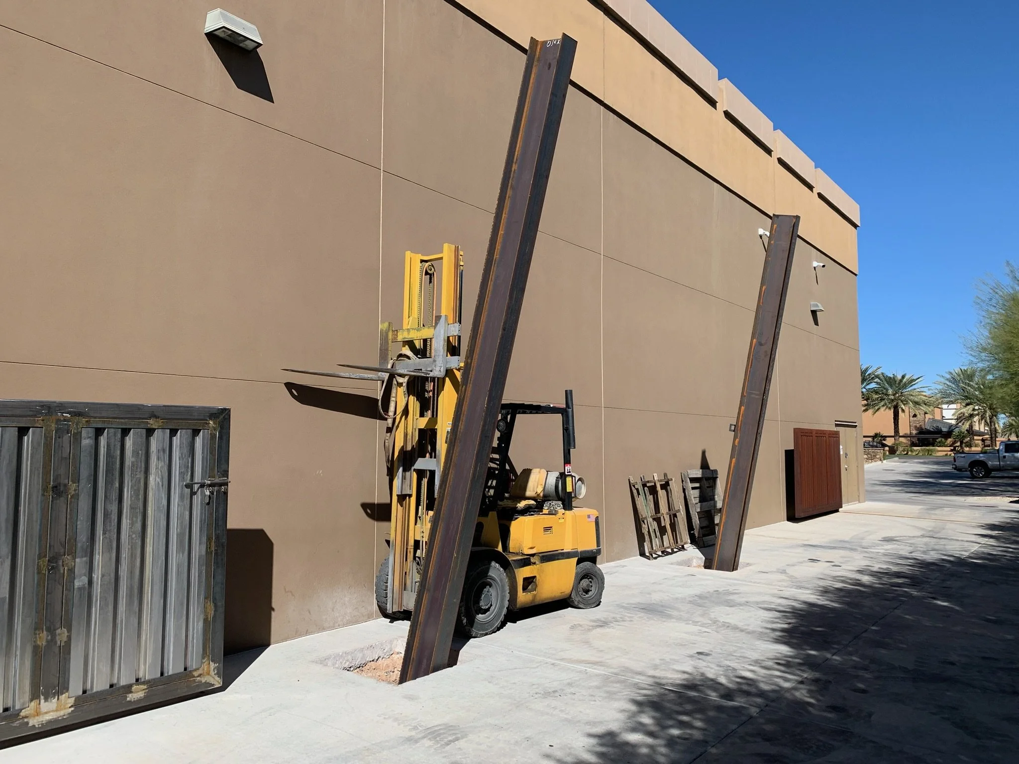 Construction site with a forklift and metal beams leaning against a beige building wall, under a clear blue sky.