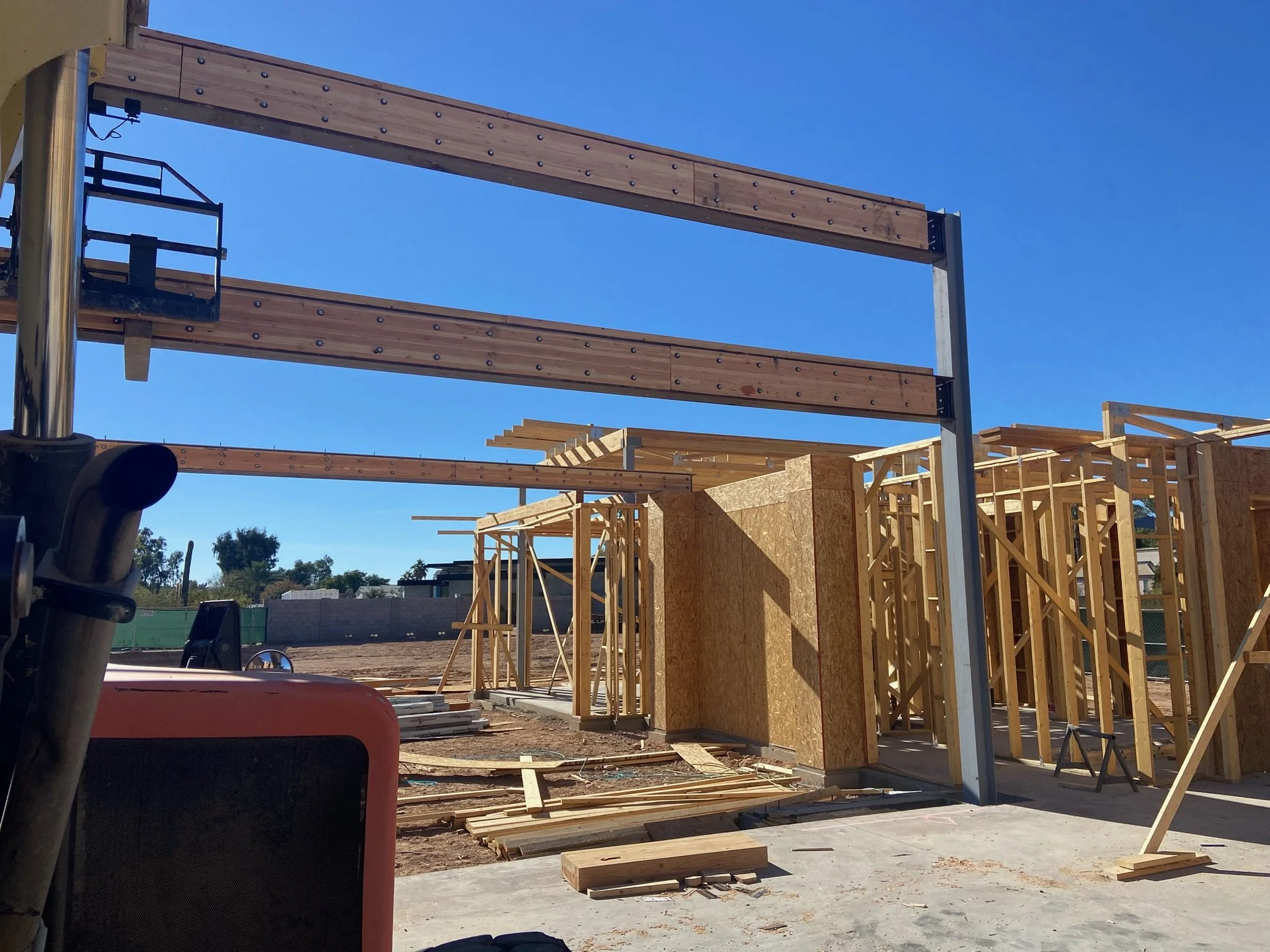 Construction site with wood framing for a building, metal beams overhead, and construction equipment under a clear blue sky.