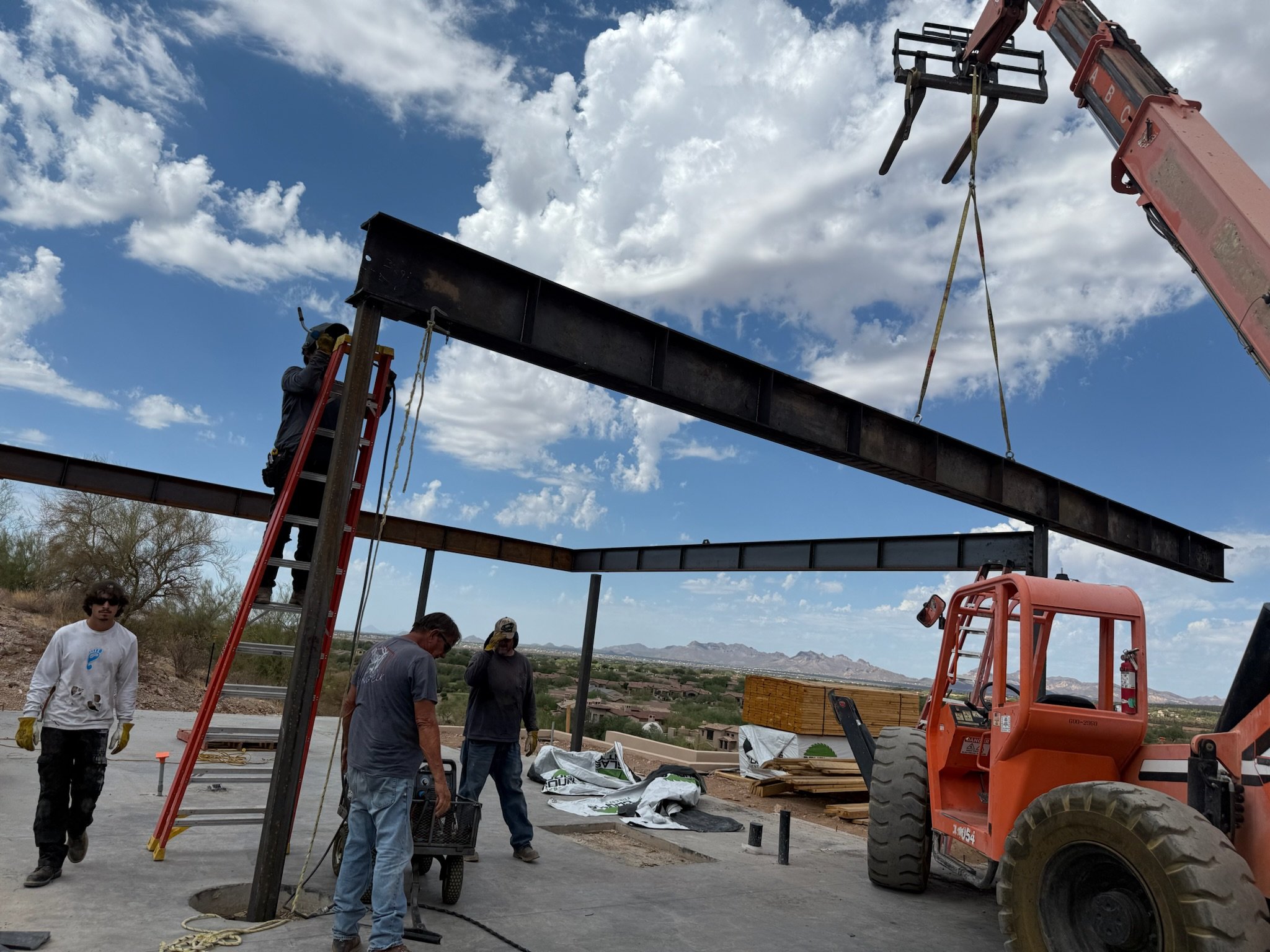 Construction site with workers and heavy machinery, assembling a steel beam structure outdoors under a partly cloudy sky.