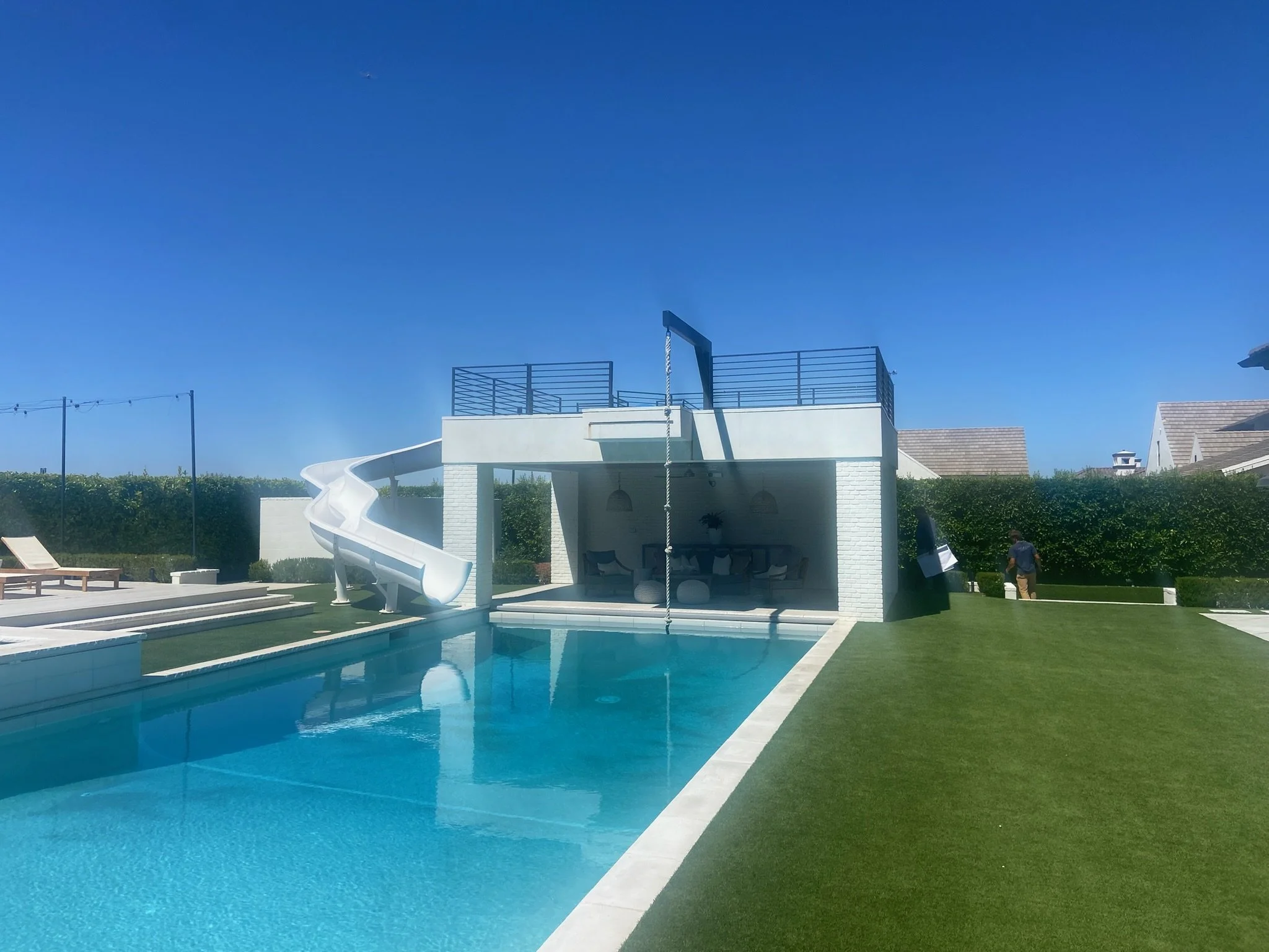 A backyard with a swimming pool, a white slide, a shaded seating area, and a small rooftop area with a ladder, surrounded by manicured grass and a hedge, with clear blue sky overhead.