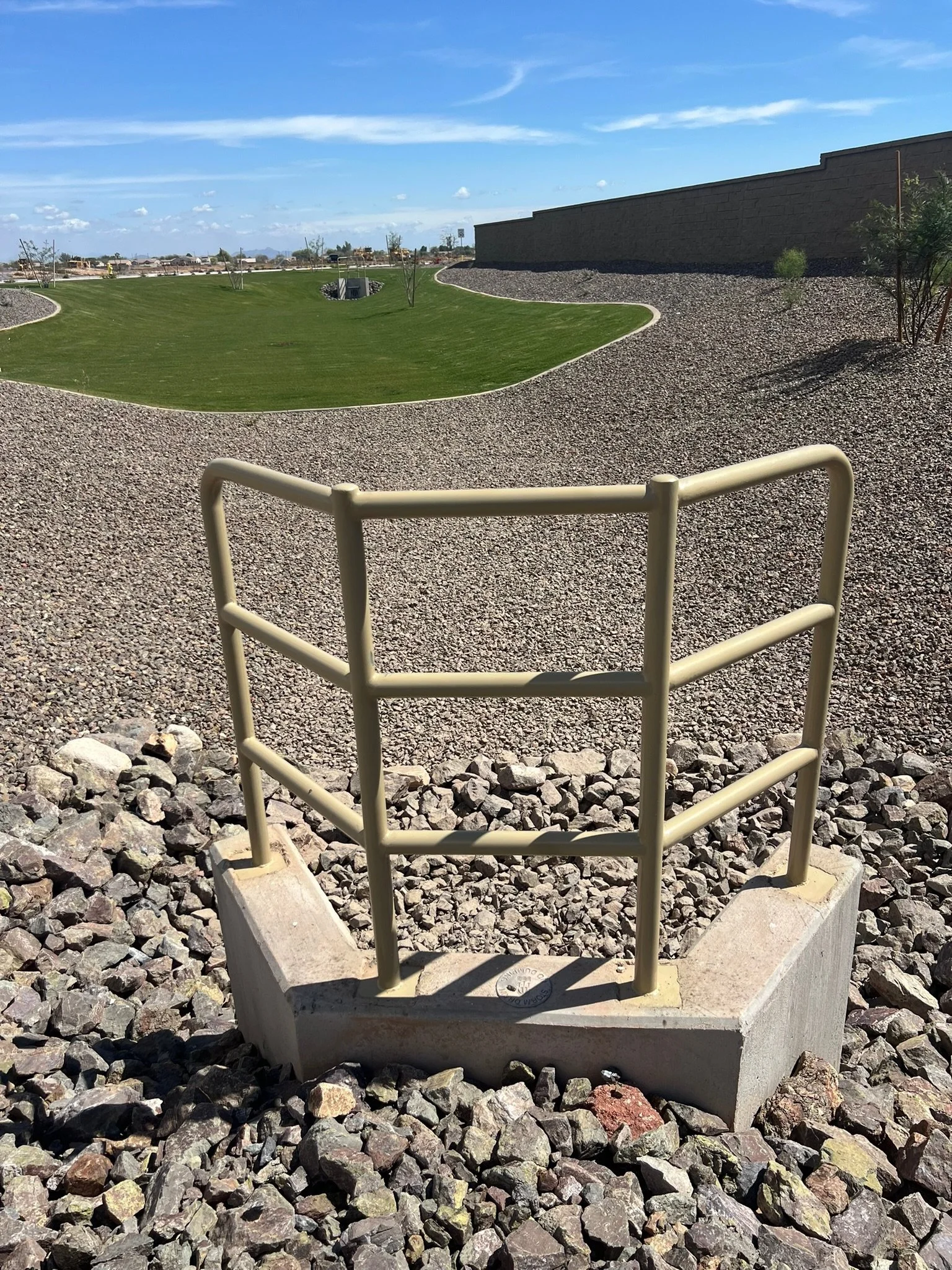 A safety railing around a concrete structure on gravel, with a grassy hill, trees, and a pool cover in the background under a blue sky with clouds.