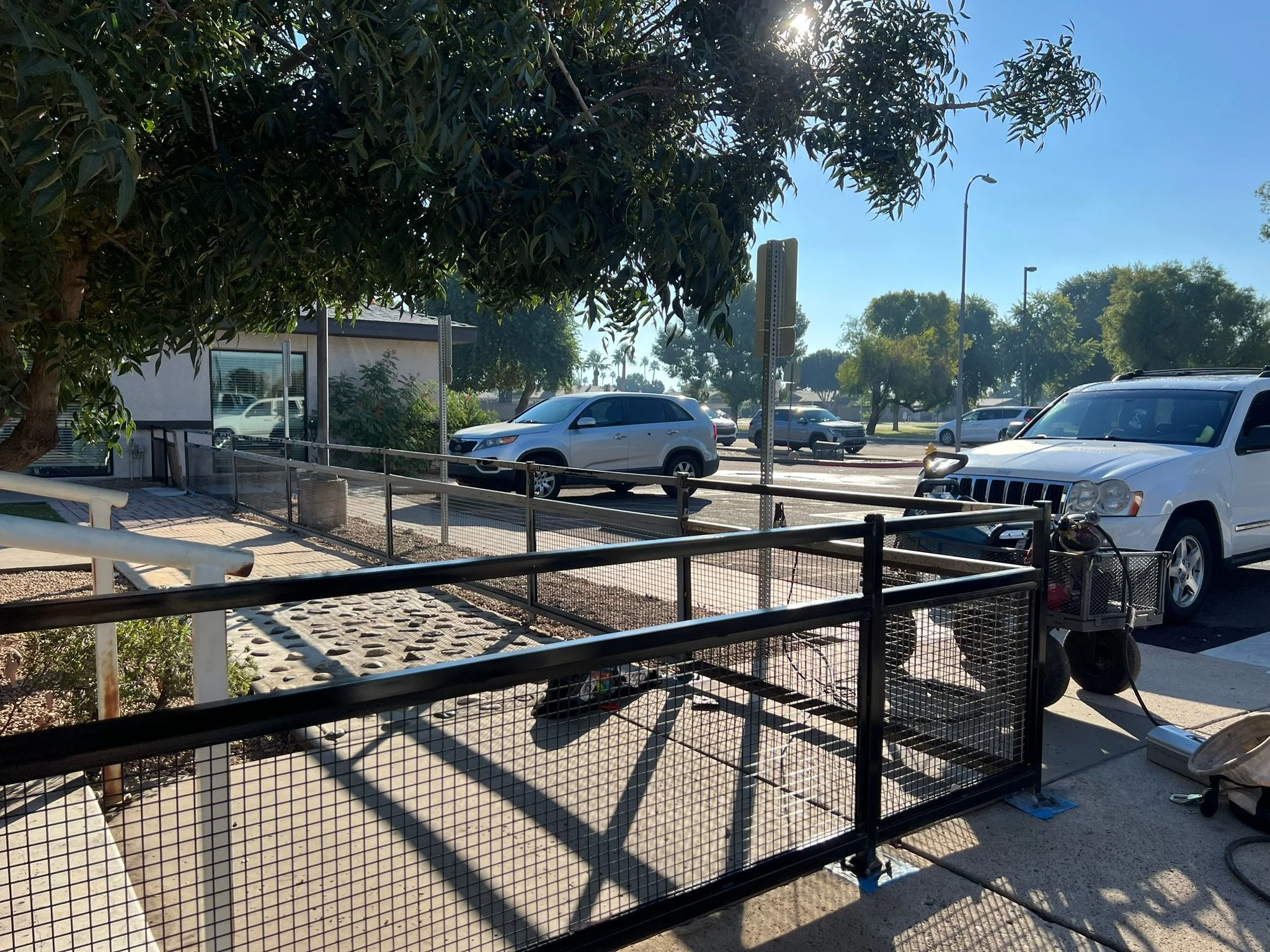 A sidewalk with a black metal fence and a tree, parked cars in a parking lot, and a street with additional vehicles and trees in the background on a sunny day.