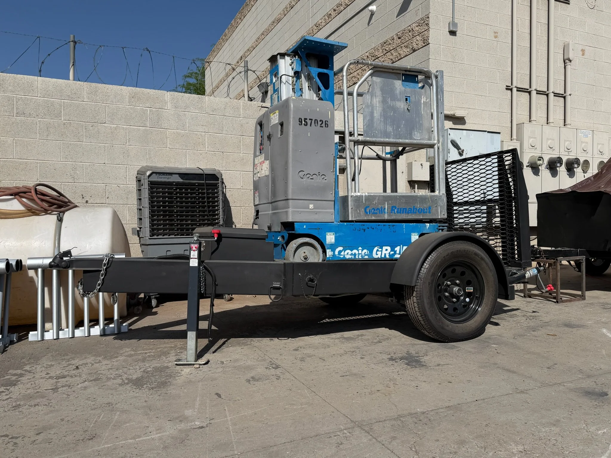A Genie GR-12 building maintenance lift parked outdoors on concrete, with a black trailer attached, nearby equipment, and a beige brick wall with electrical boxes and barbed wire.