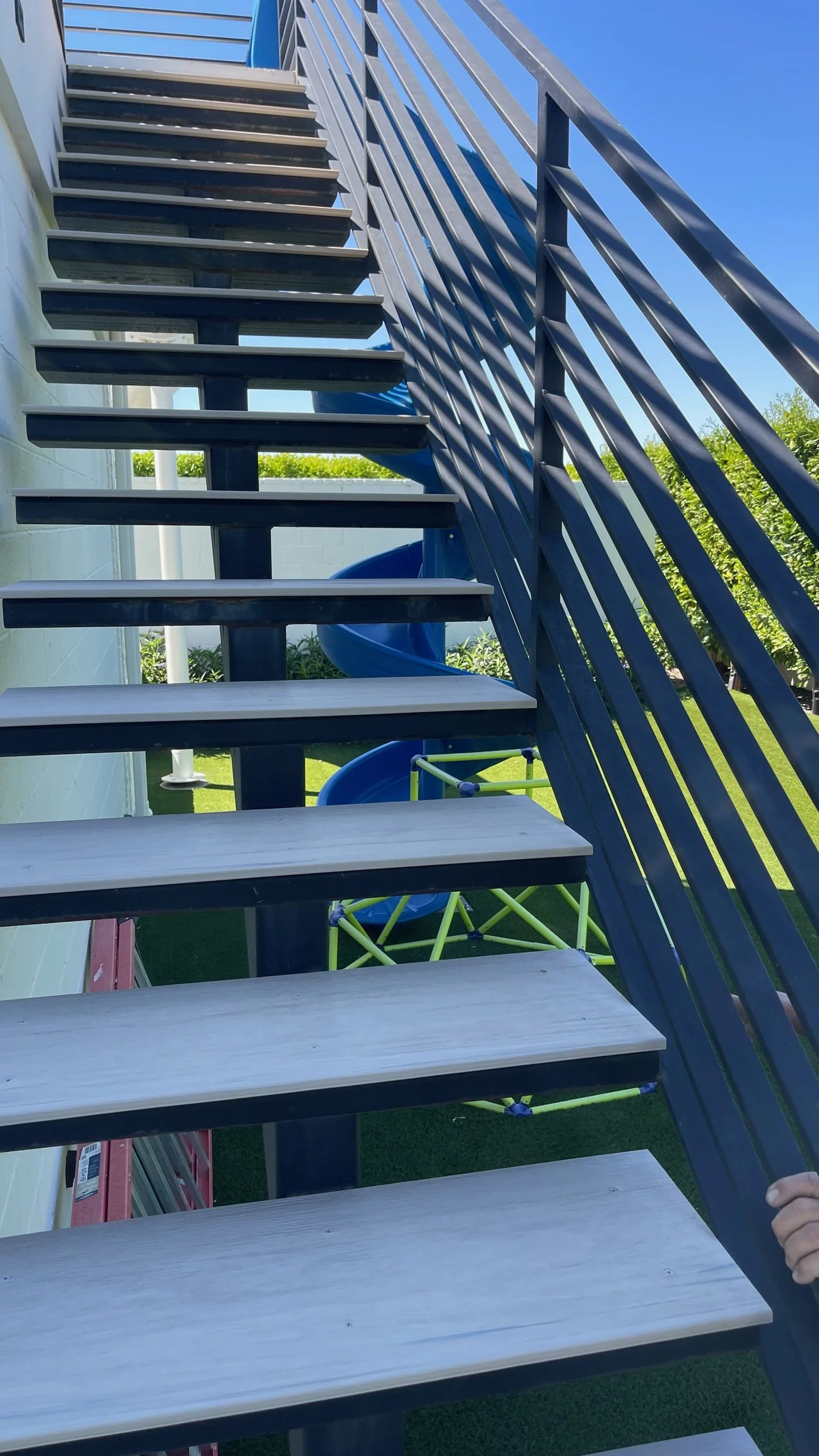 Outdoor staircase with black metal railing and wooden steps, leading up to a higher level. Below the staircase, there is a blue spiral slide and a yellow climbing structure. The background includes a white wall, green grass, and a clear blue sky.
