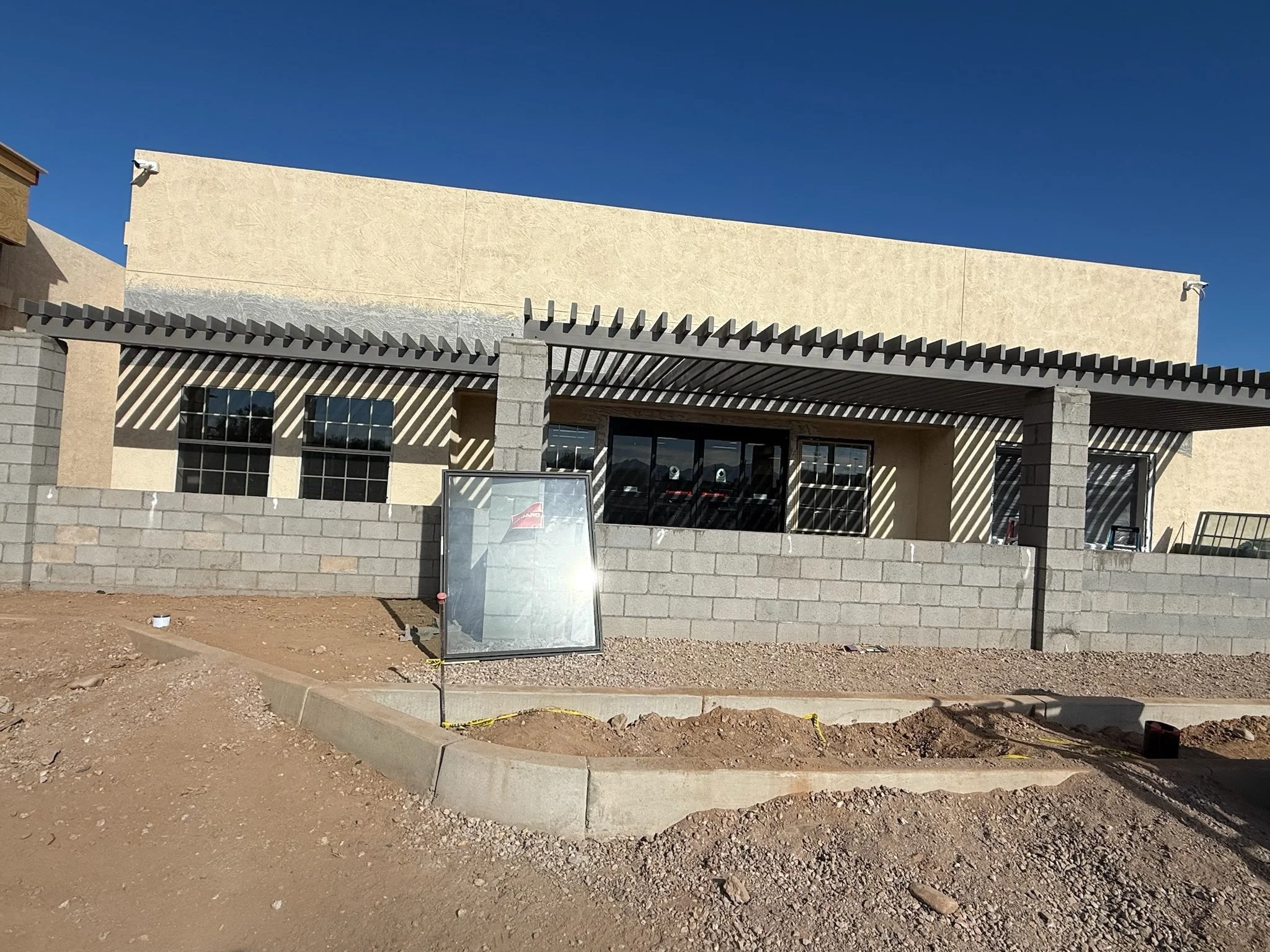 Construction site of a building with a gray concrete block wall, beige stucco exterior, and dark window frames. The area in front is dirt with some construction materials and a partially assembled sign.