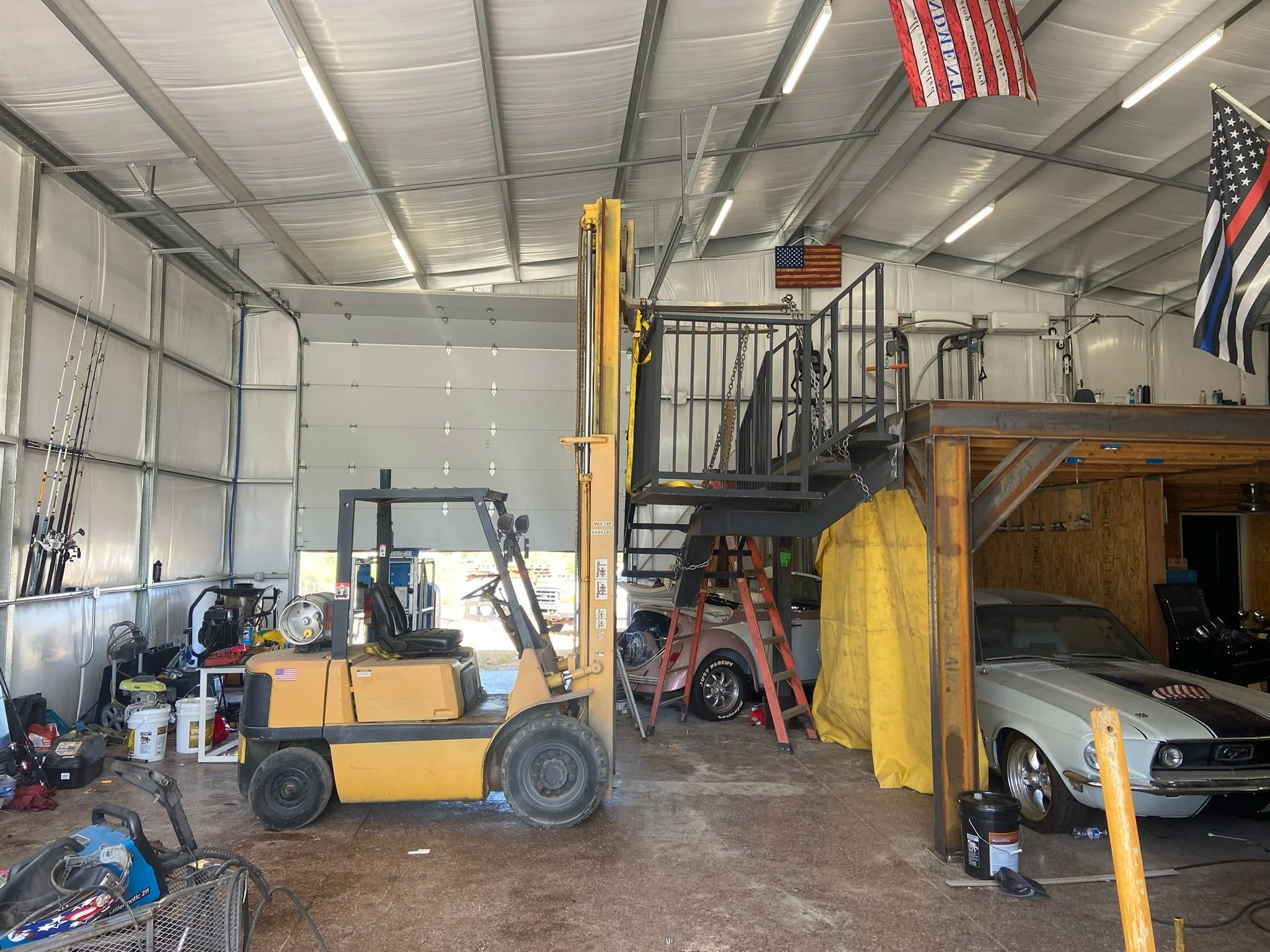 A garage interior with a yellow forklift, vintage cars, and tools, featuring American flags and a black and white with a blue stripe flag.