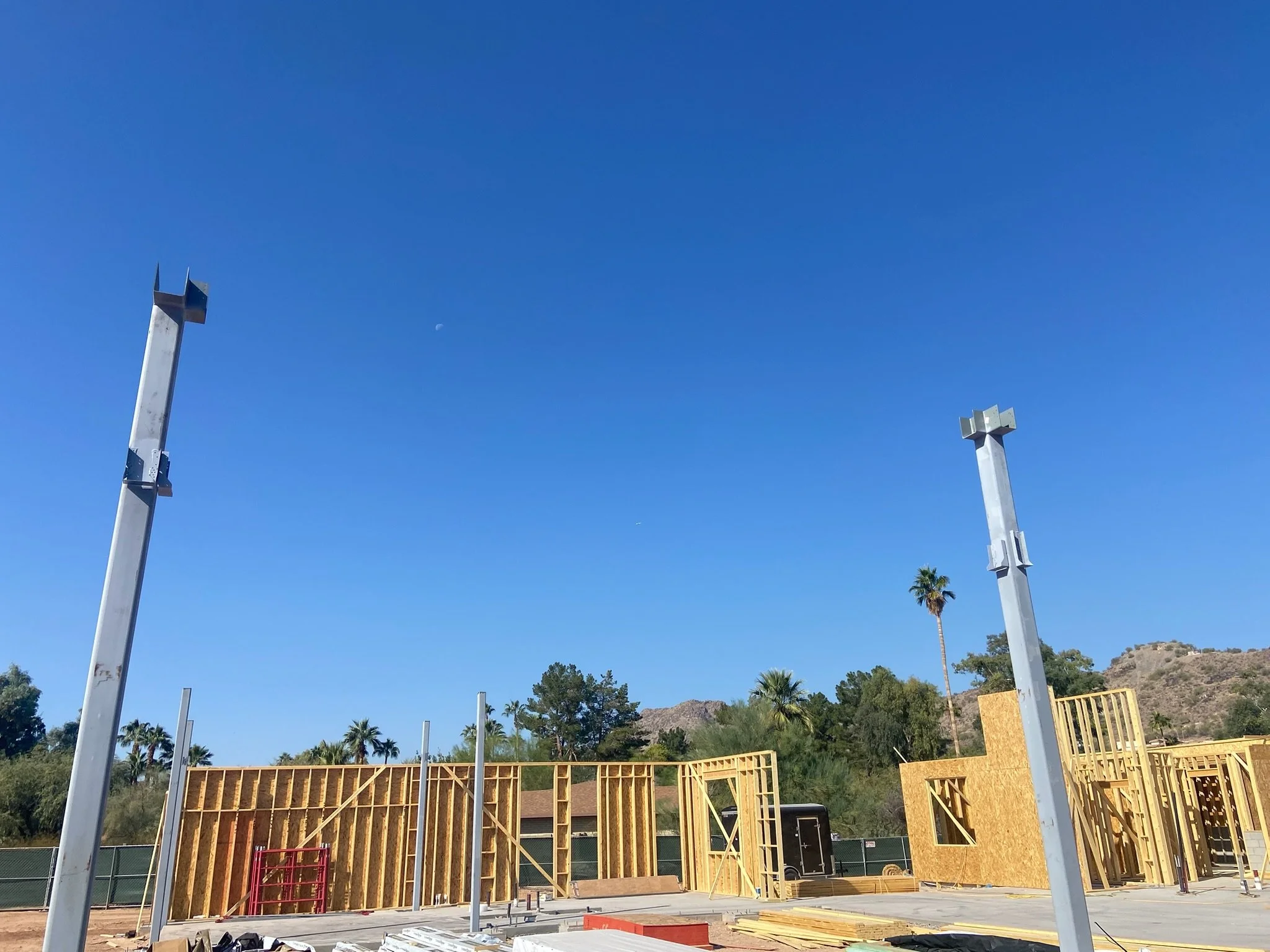 Construction site with wooden frames for walls, metal beams, and a blue sky with a faint moon, in a hilly area with palm trees.