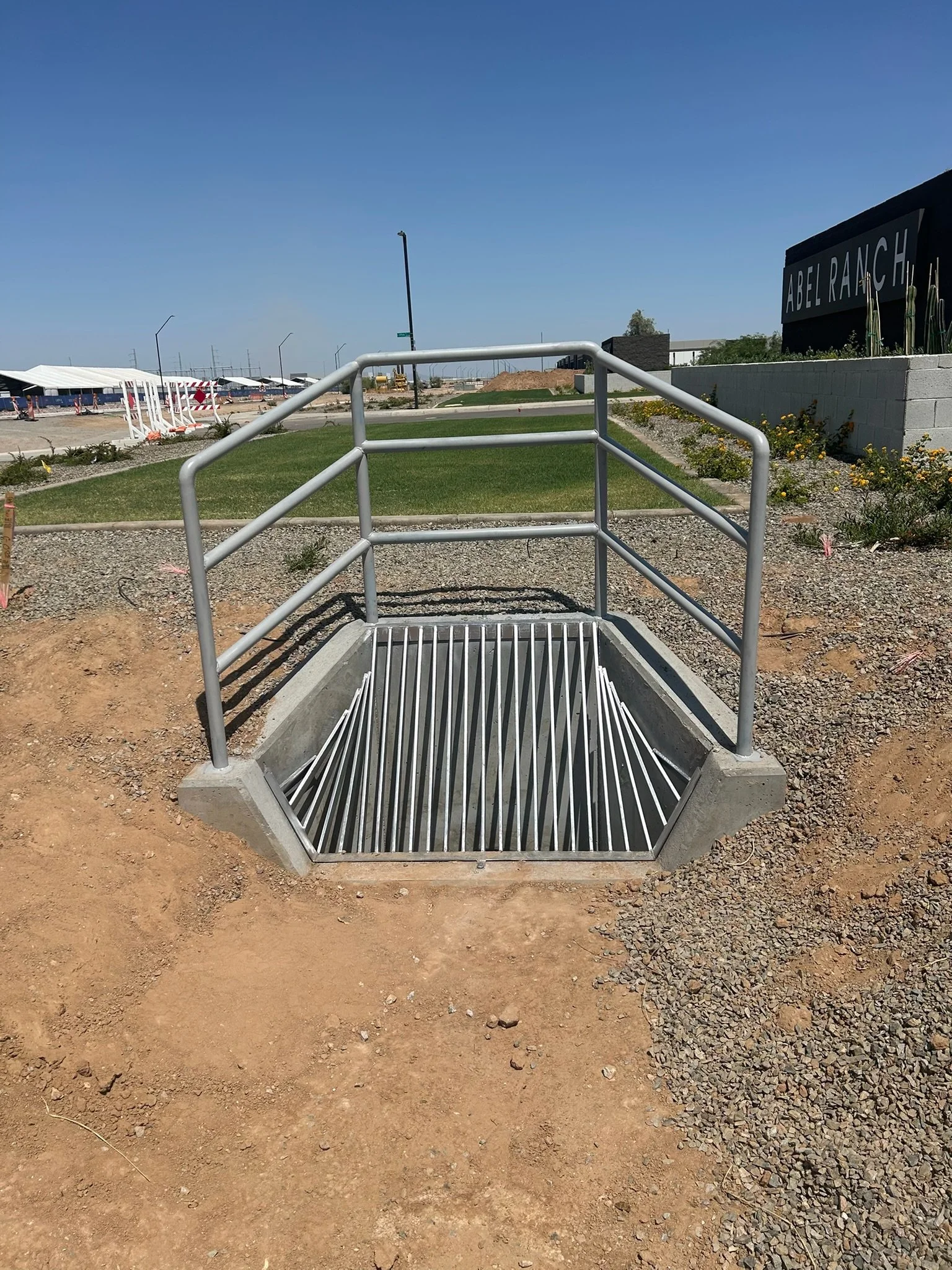 Underground utility access hatch with metal grate and railing, installed in a landscaped area with grass and flowers, under a clear blue sky.
