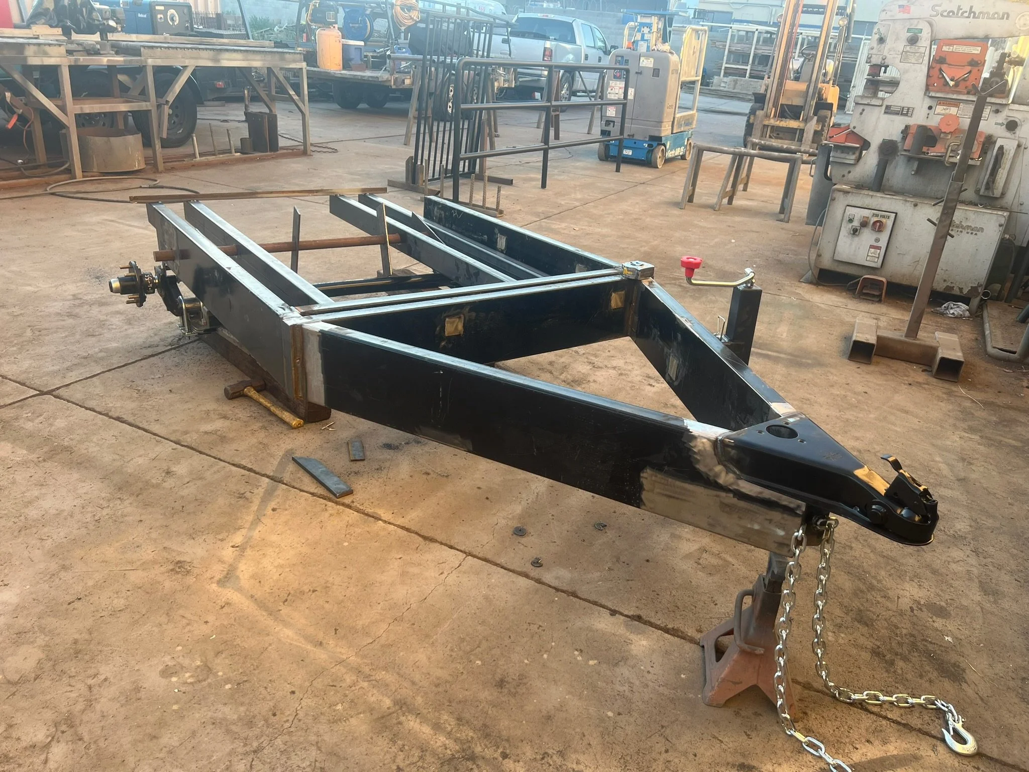 Black metal trailer frame resting on garage floor with chains attached to front, in a workshop with various tools, machinery, and vehicles in the background.