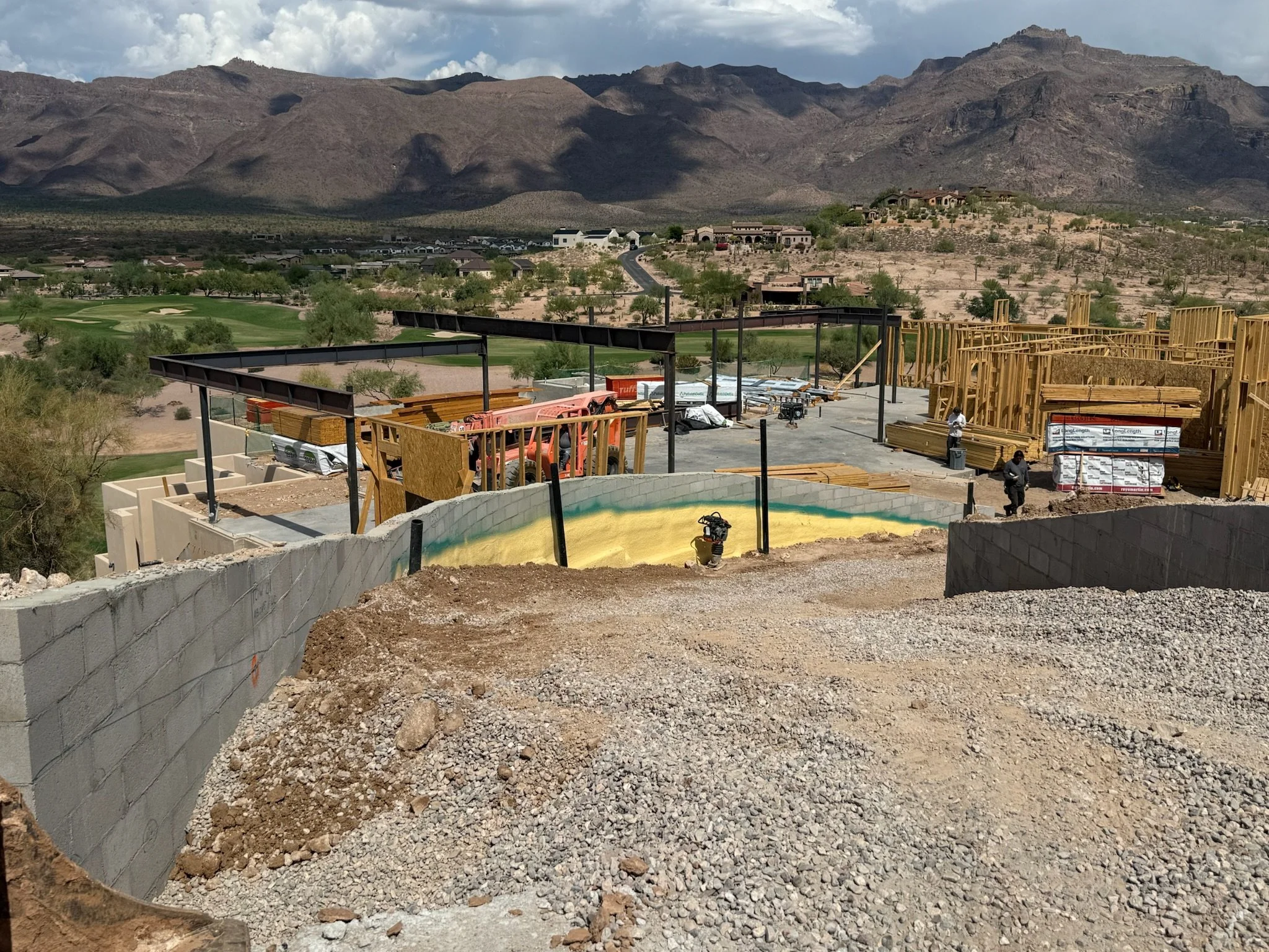 Construction site on a hillside with framing, equipment, and workers, overlooking a desert landscape with mountains and houses in the distance.