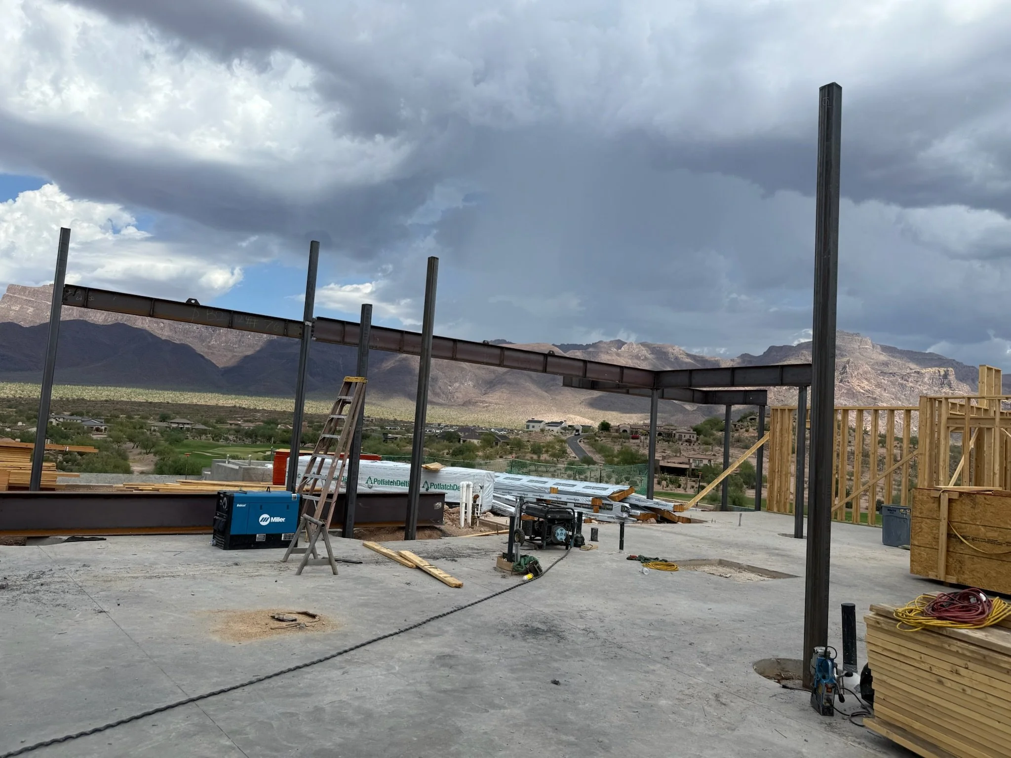 Construction site with steel framework partially built, tools, wooden planks, and a ladder, set against a mountainous desert landscape with cloudy skies.