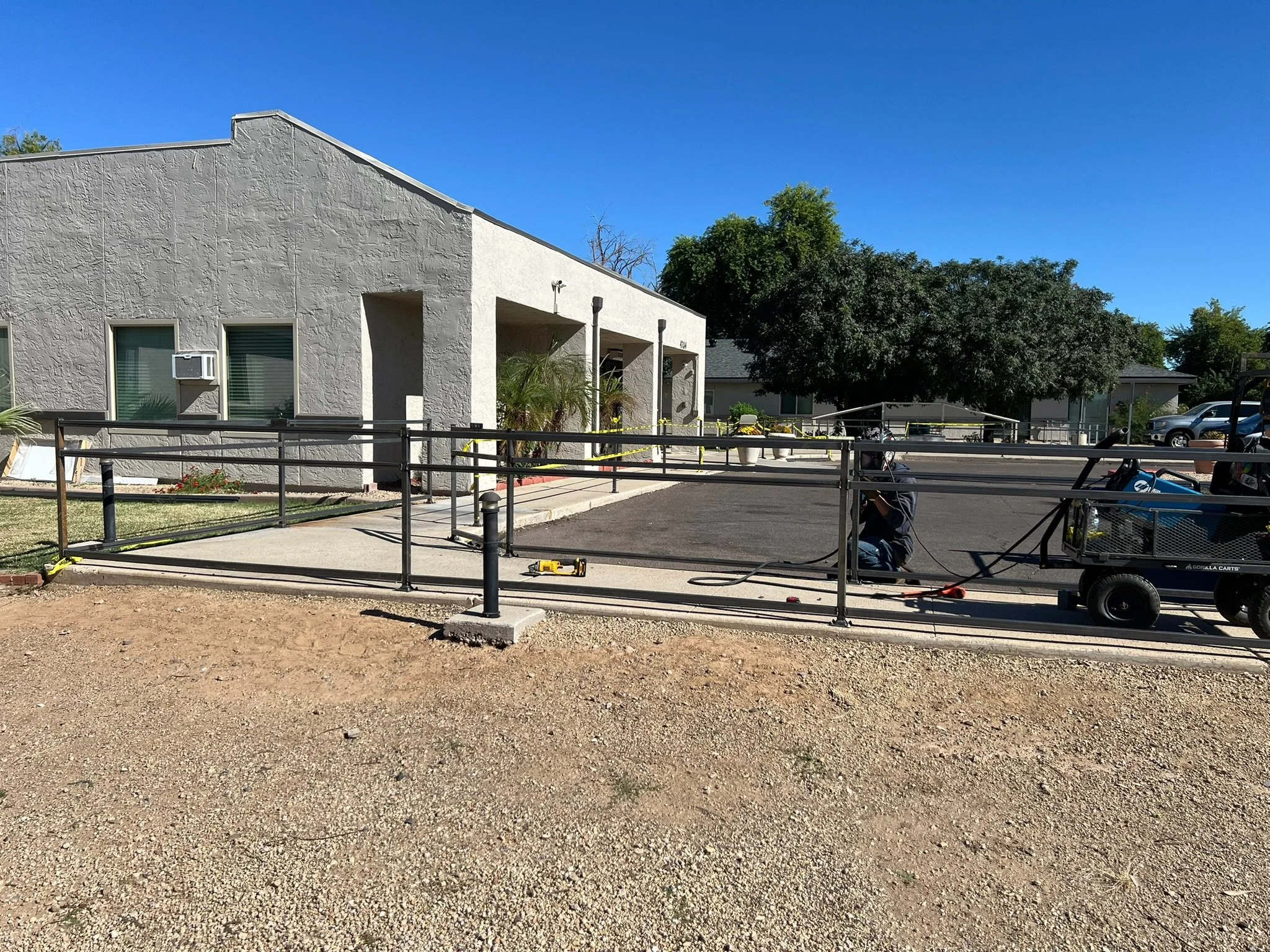 Construction workers installing a ramp in front of a beige residential building on a sunny day. The ramp is surrounded by metal railings, with tools and equipment nearby. There are trees and parked cars in the background.