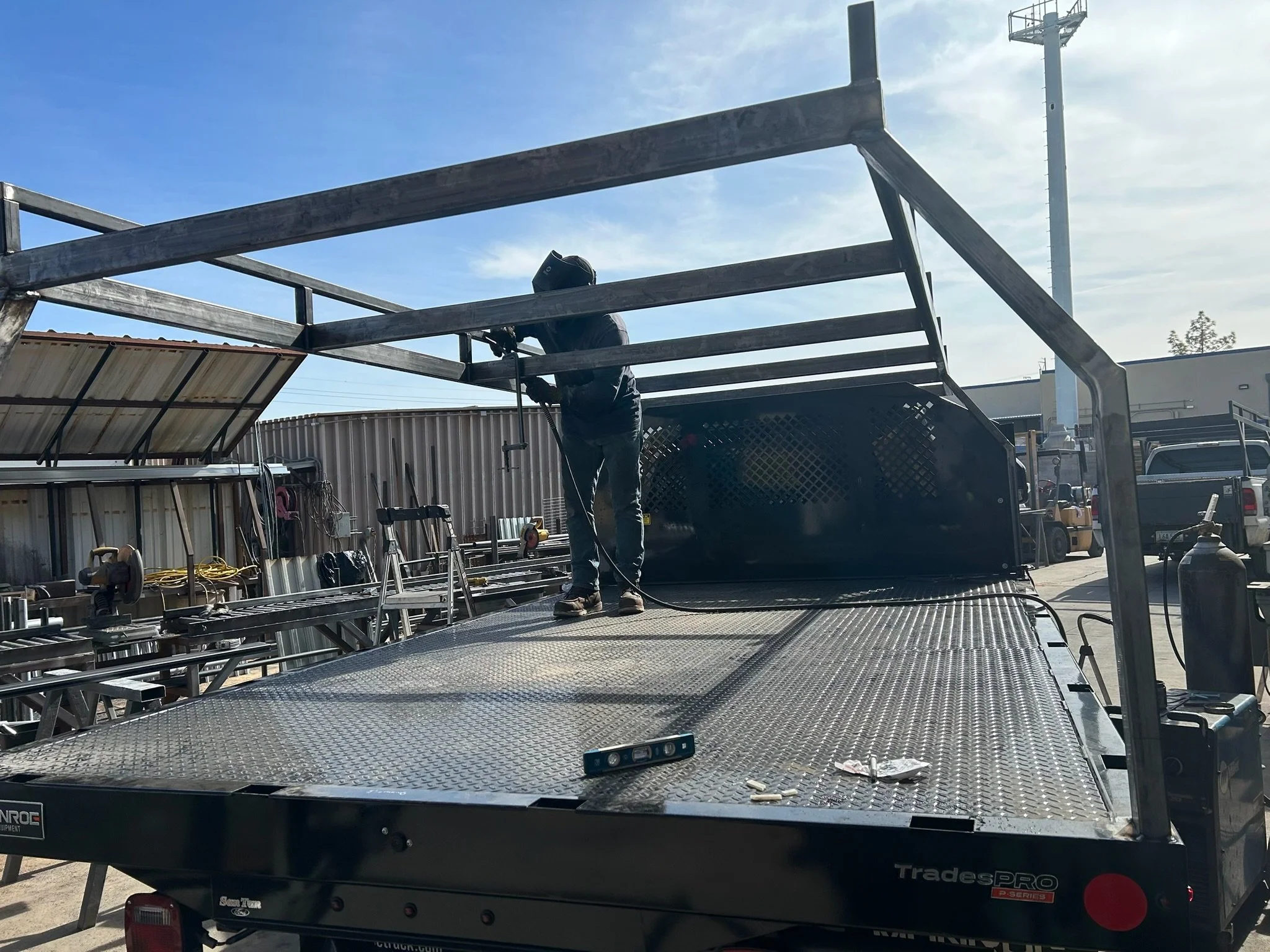 A worker welding on a flatbed trailer in an industrial yard, with various tools and equipment visible in the background, and a clear blue sky overhead.