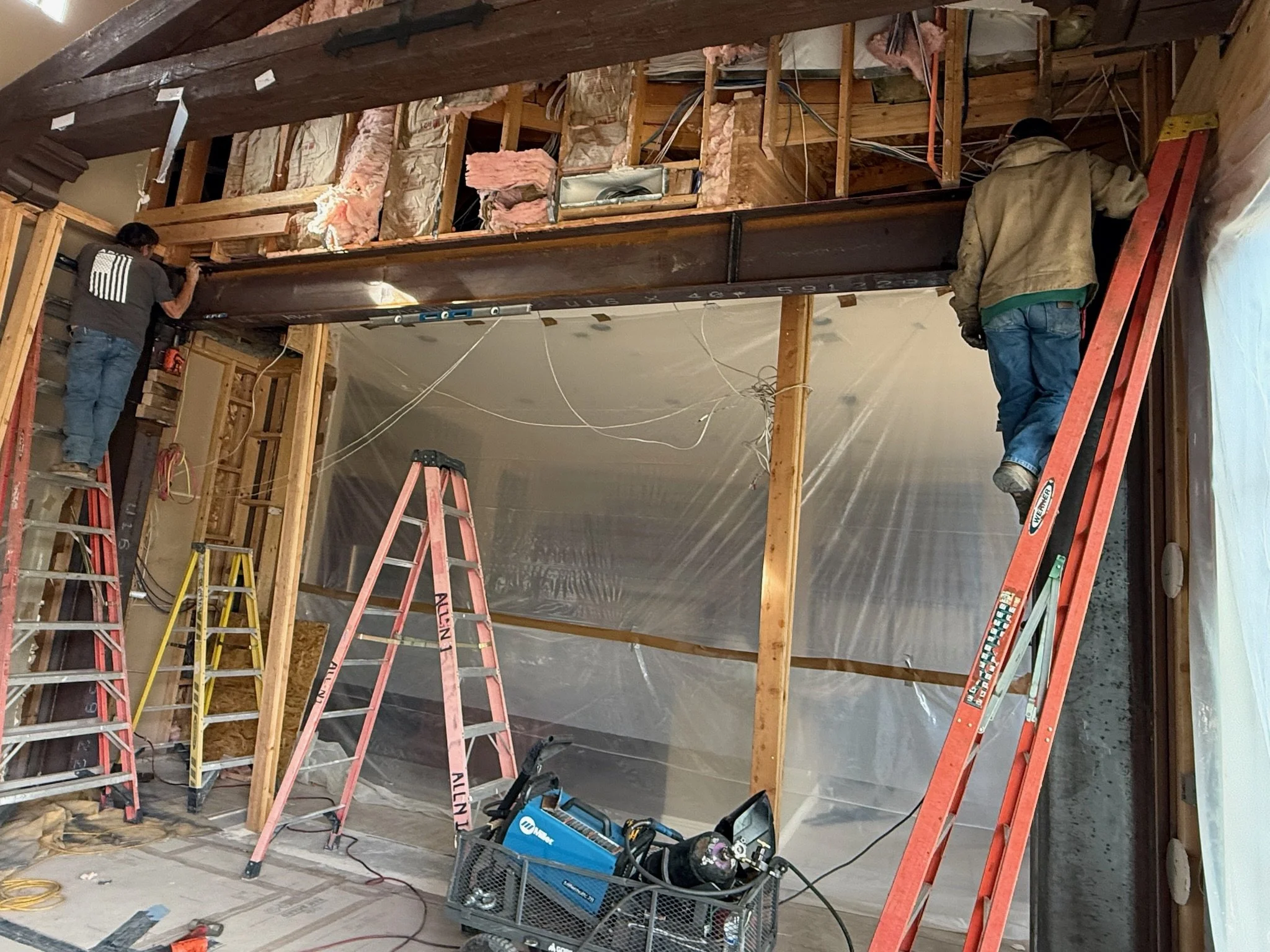 Two construction workers are working inside a building, standing on ladders, installing or repairing a large steel beam. The space is under renovation with exposed wooden framing, insulation, and wiring.
