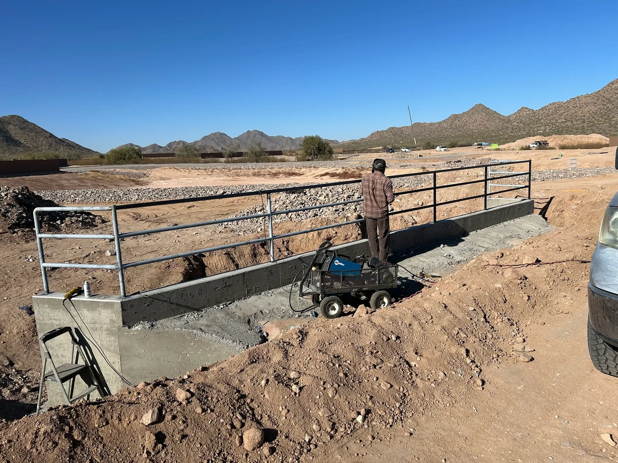 A construction worker standing behind a safety railing on a concrete structure in a desert landscape with mountains in the background.
