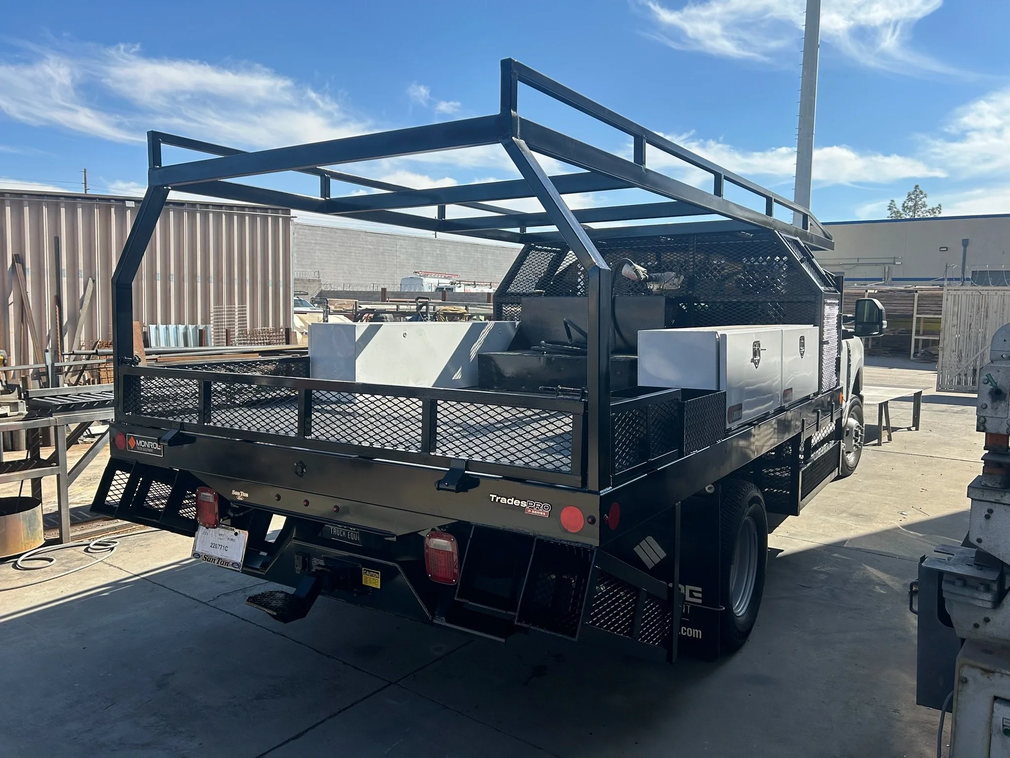 Black flatbed utility truck with a metal frame on top, parked in an outdoor industrial lot, with a concrete ground and a partly cloudy sky.